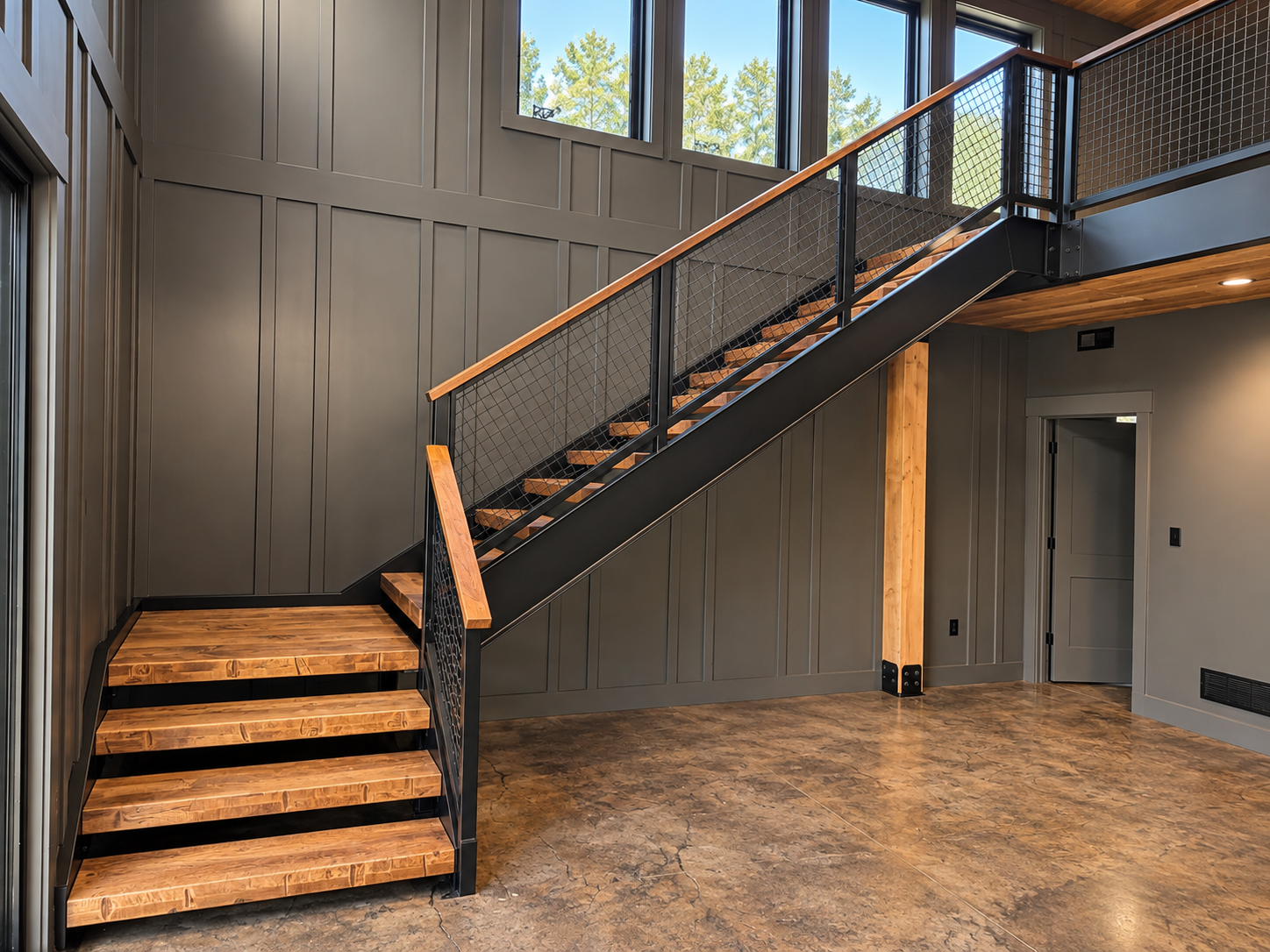 Modern interior with wooden staircase and metal railings, gray wall paneling, and large windows showing green trees outside.