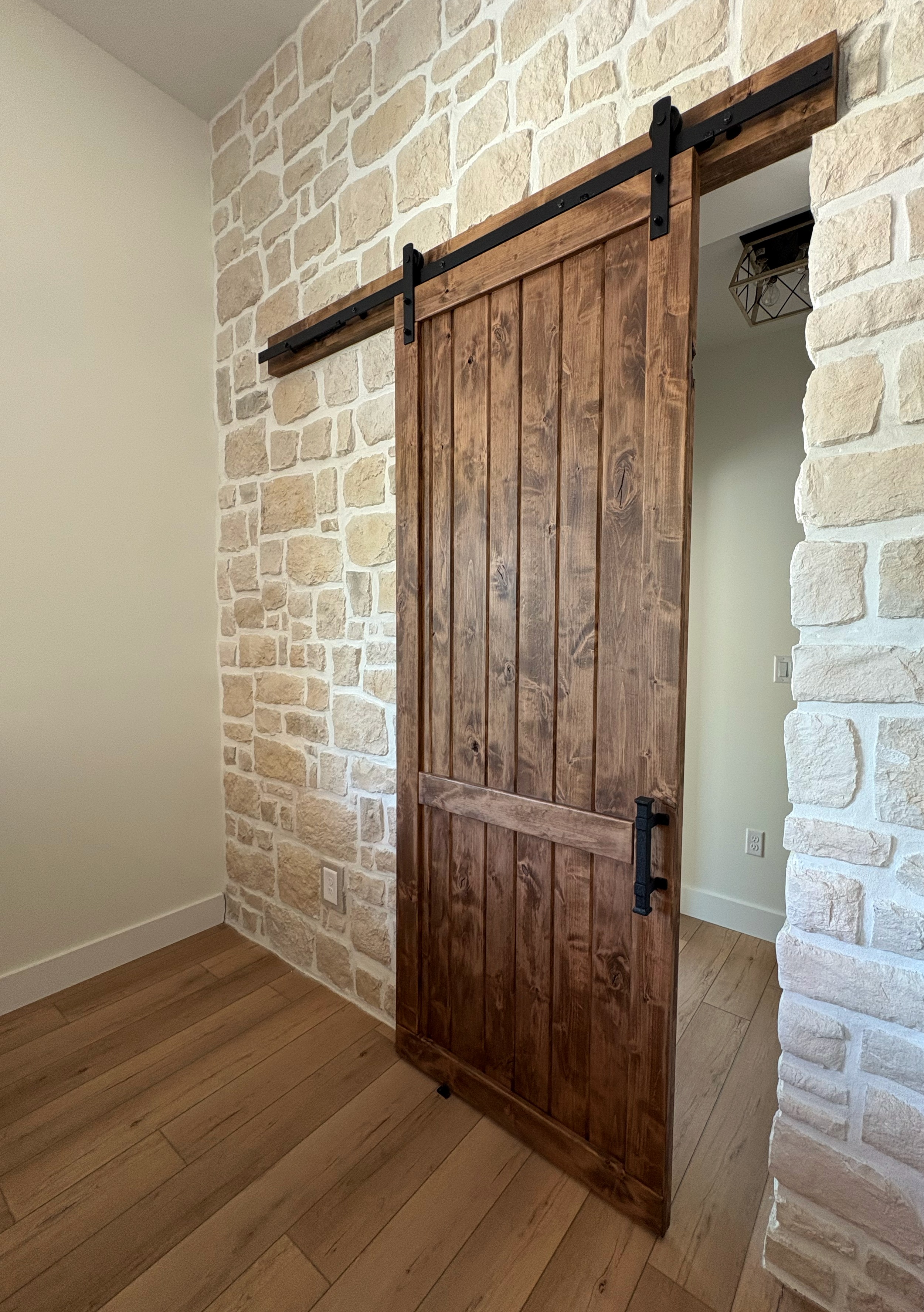 A wooden sliding barn door on a black metal track, partially open, installed on a beige brick wall in a room with hardwood flooring.