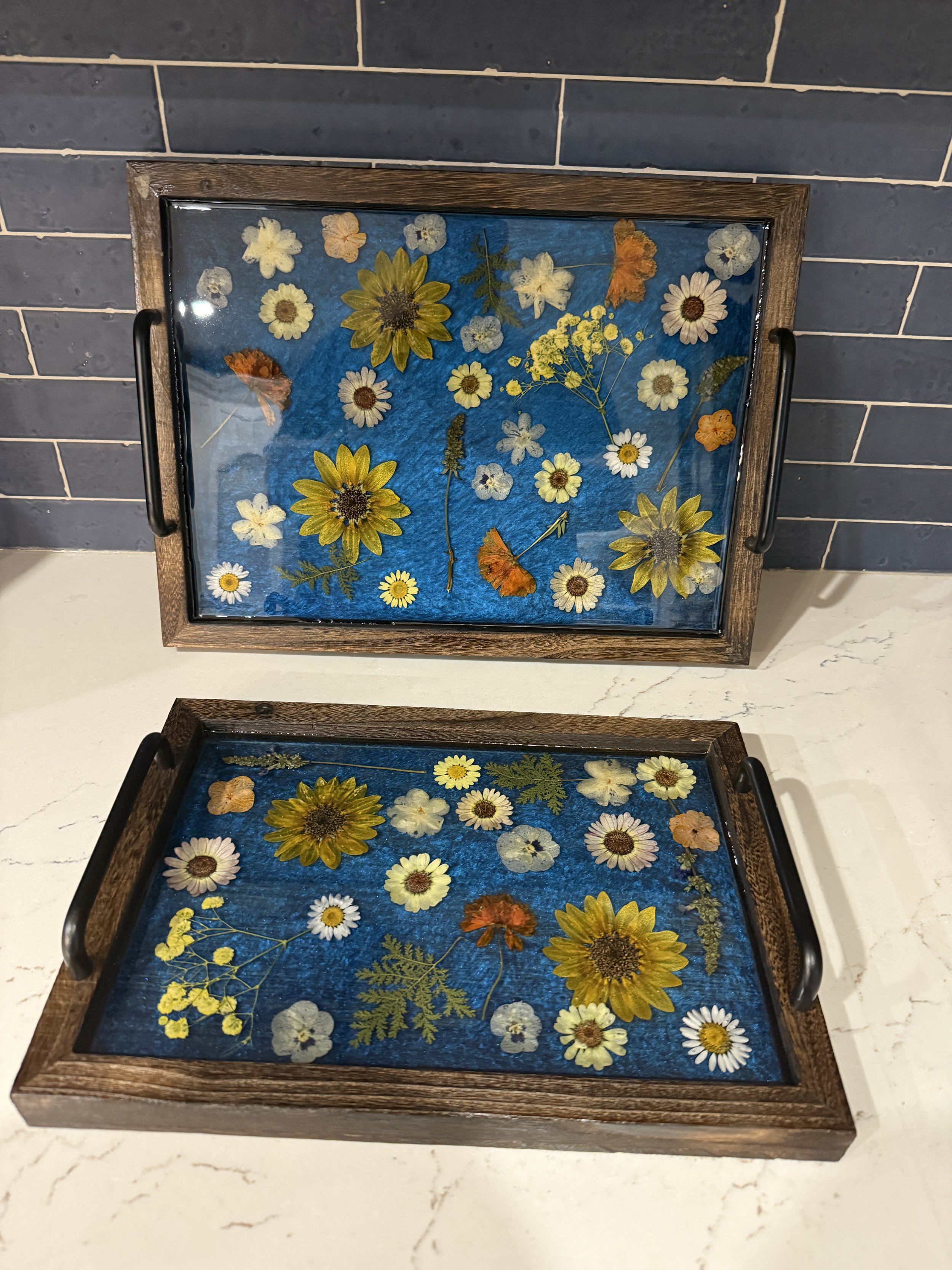 Two wooden trays with glass tops containing pressed flowers and leaves, placed on a white marble surface. The trays have black handles, and the pressed flowers include daisies, sunflowers, and other small wildflowers on a blue background.