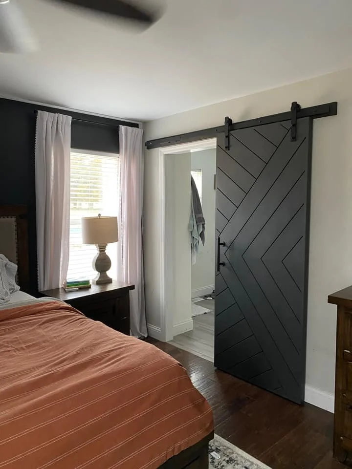 Interior of a bedroom with a sliding barn door, a window with white curtains, a nightstand with a lamp, and part of a bed with a peach-colored blanket.