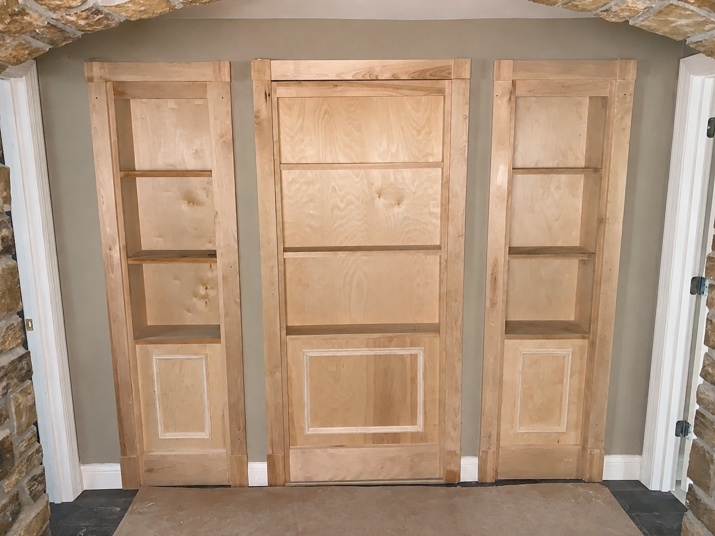 Three unfinished wooden doors with shelving, one in the middle flanked by two on either side, installed on a beige wall with stone accents on the sides.