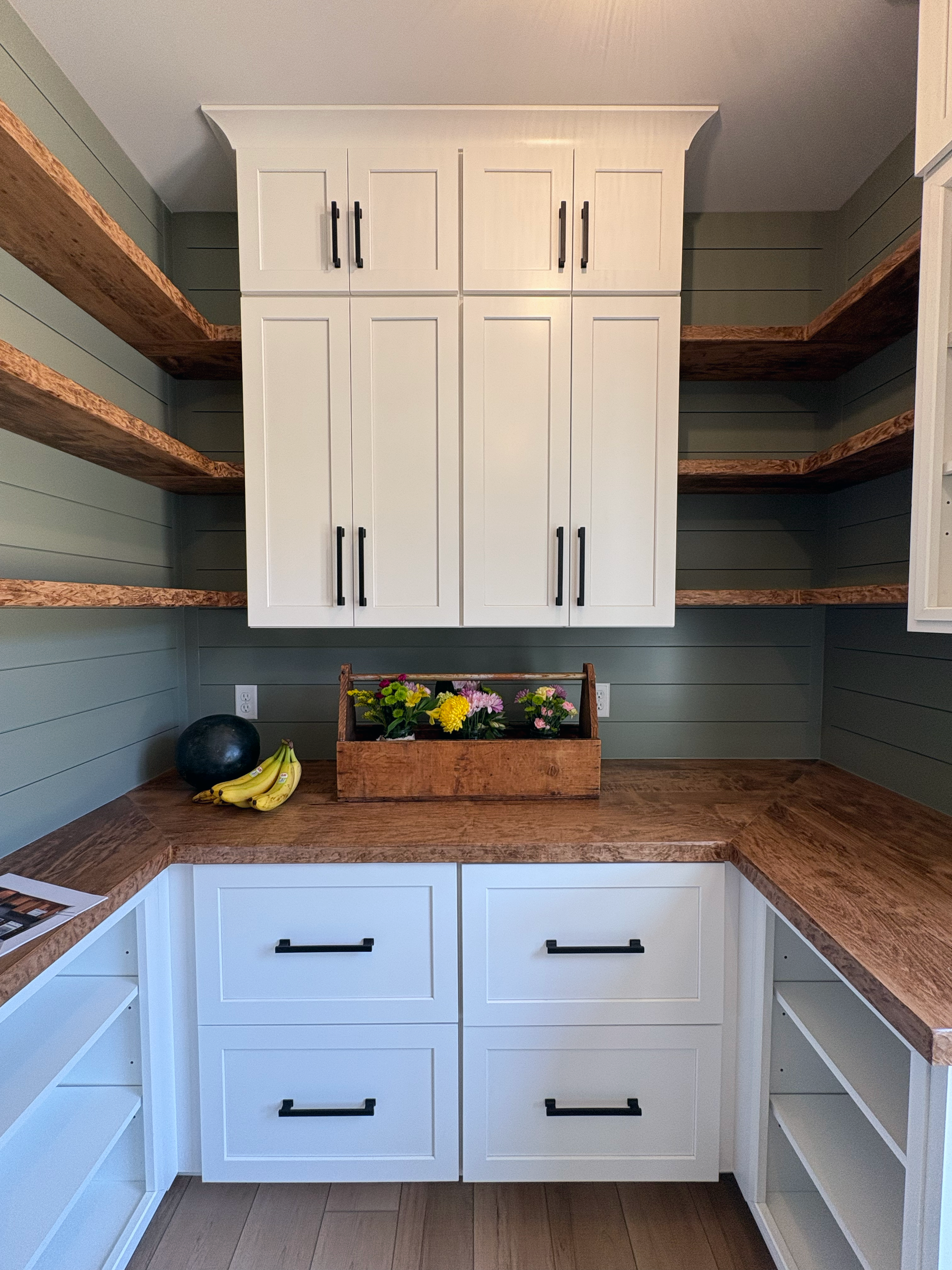 A kitchen corner with white cabinets, wooden countertops, open shelves, a bowl of bananas, a black ball, a wooden box with colorful flowers, and a paper on the counter.