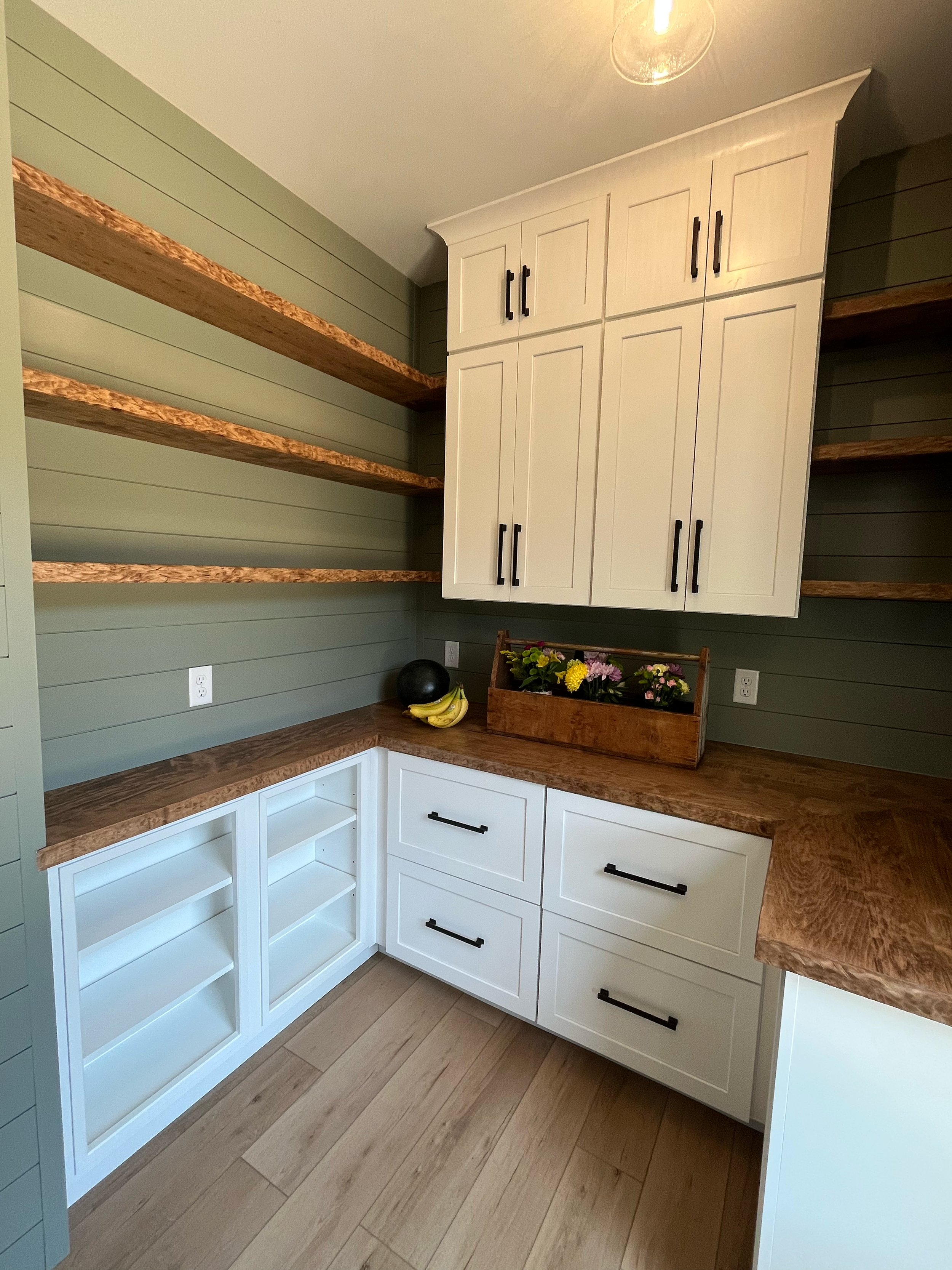 A kitchen corner with white cabinets, black handles, and open white shelves. Green shiplap walls, wooden countertops, a black vase, and a banana bunch with flowers in a wooden crate.