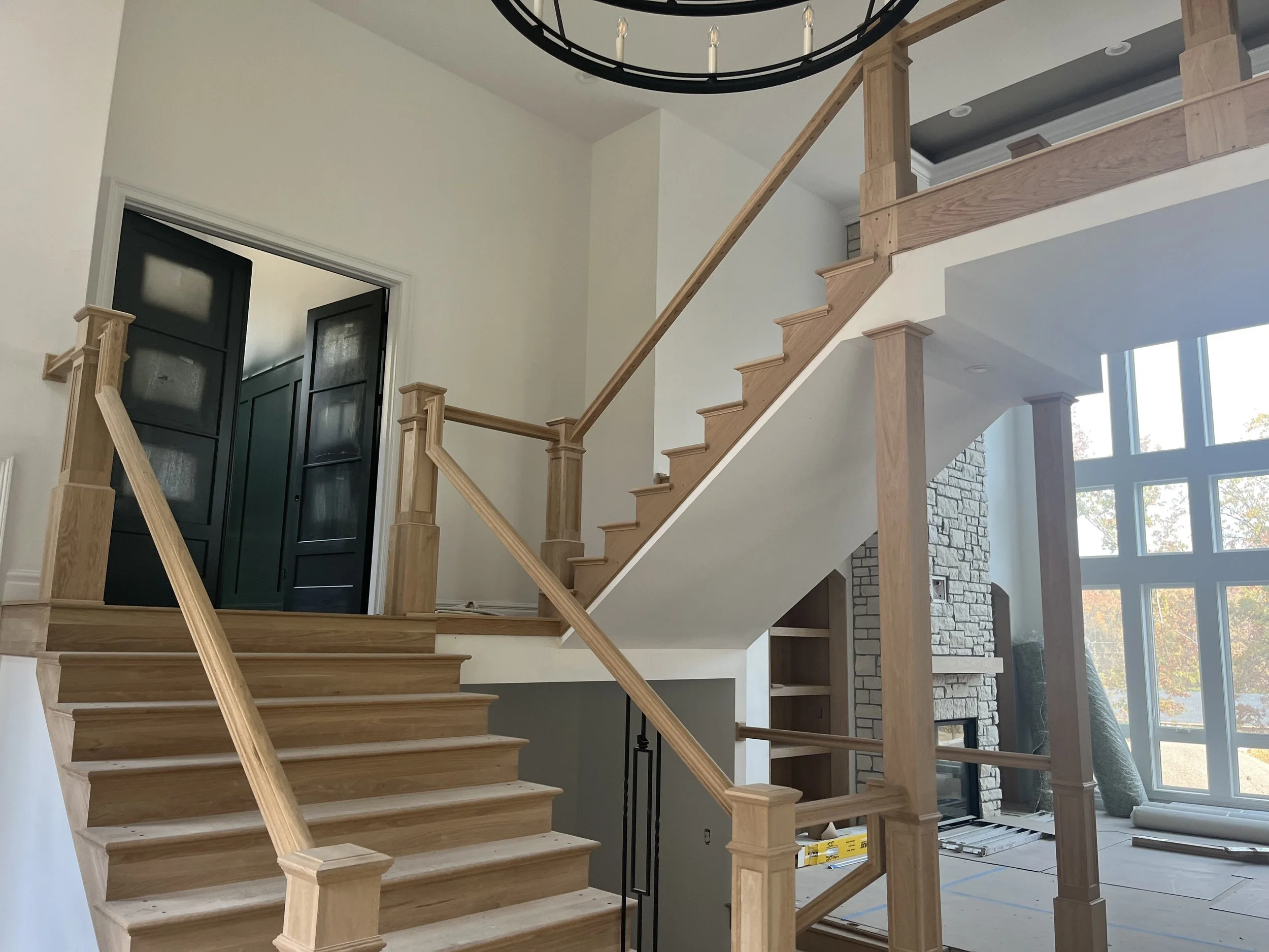 Interior view of a house under construction, featuring a partially built wooden staircase, large windows, and a view of an unfinished room with brick fireplace and construction materials.