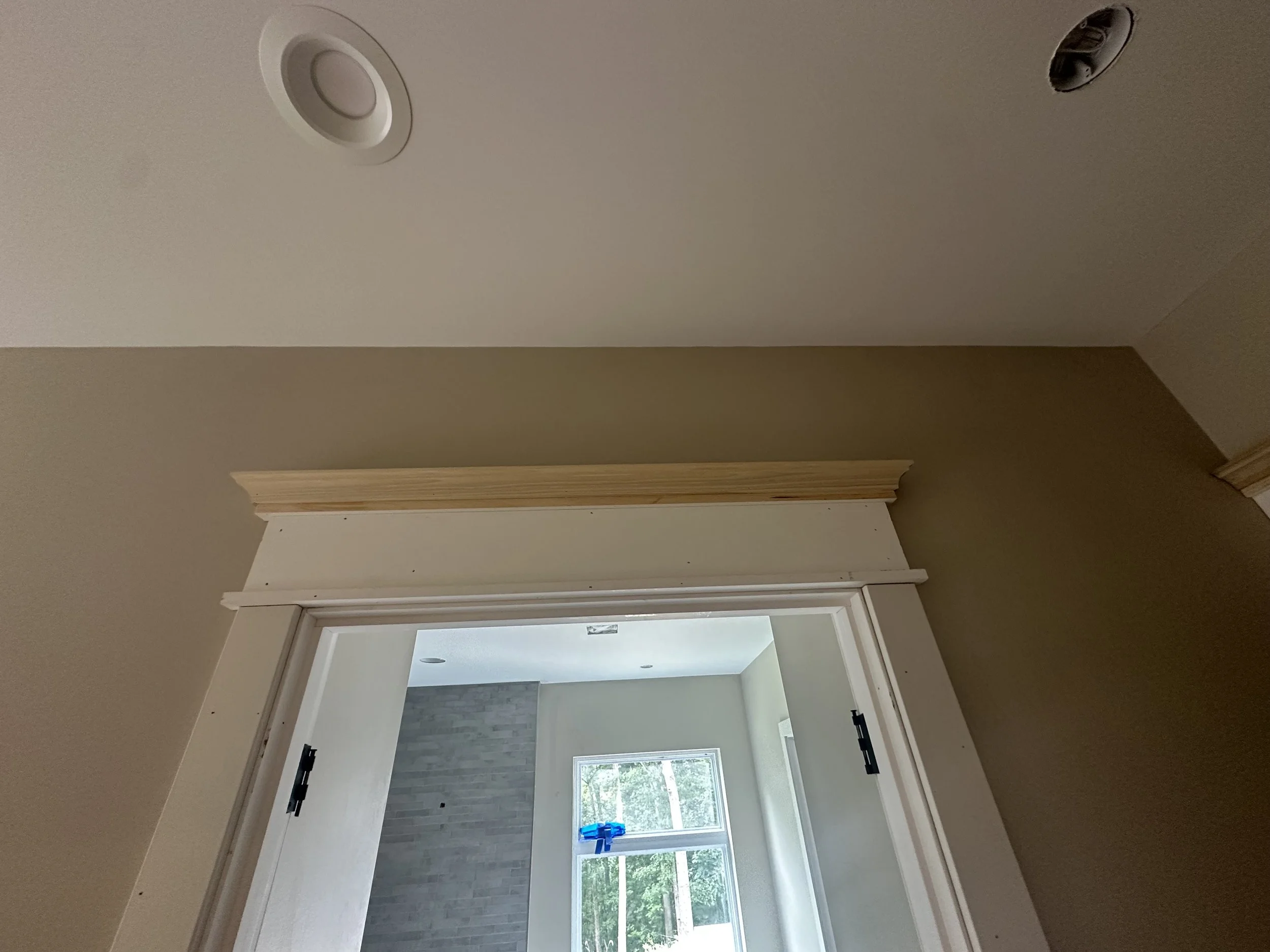 Interior view of a room with a door frame leading to a space with a window, a brick wall, and a blue decorative item. The ceiling has a recessed light and a smoke detector.