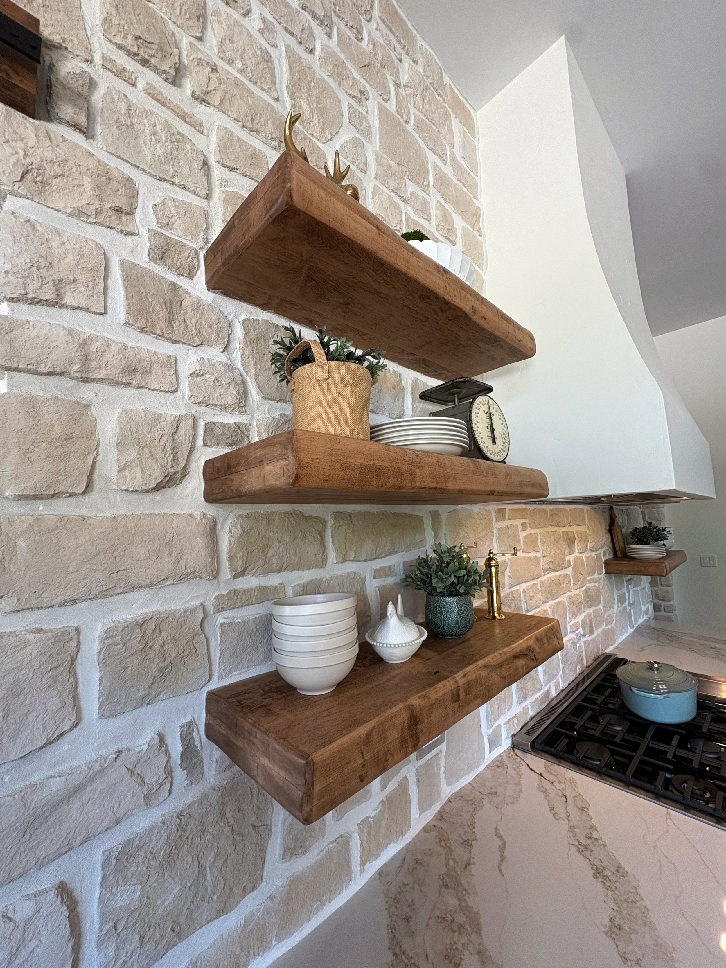 Kitchen with a brick wall, three wooden shelves holding decorative items including vases, bowls, a scale, and small plants, and a gas cooktop with a blue pot on it, marble countertop.