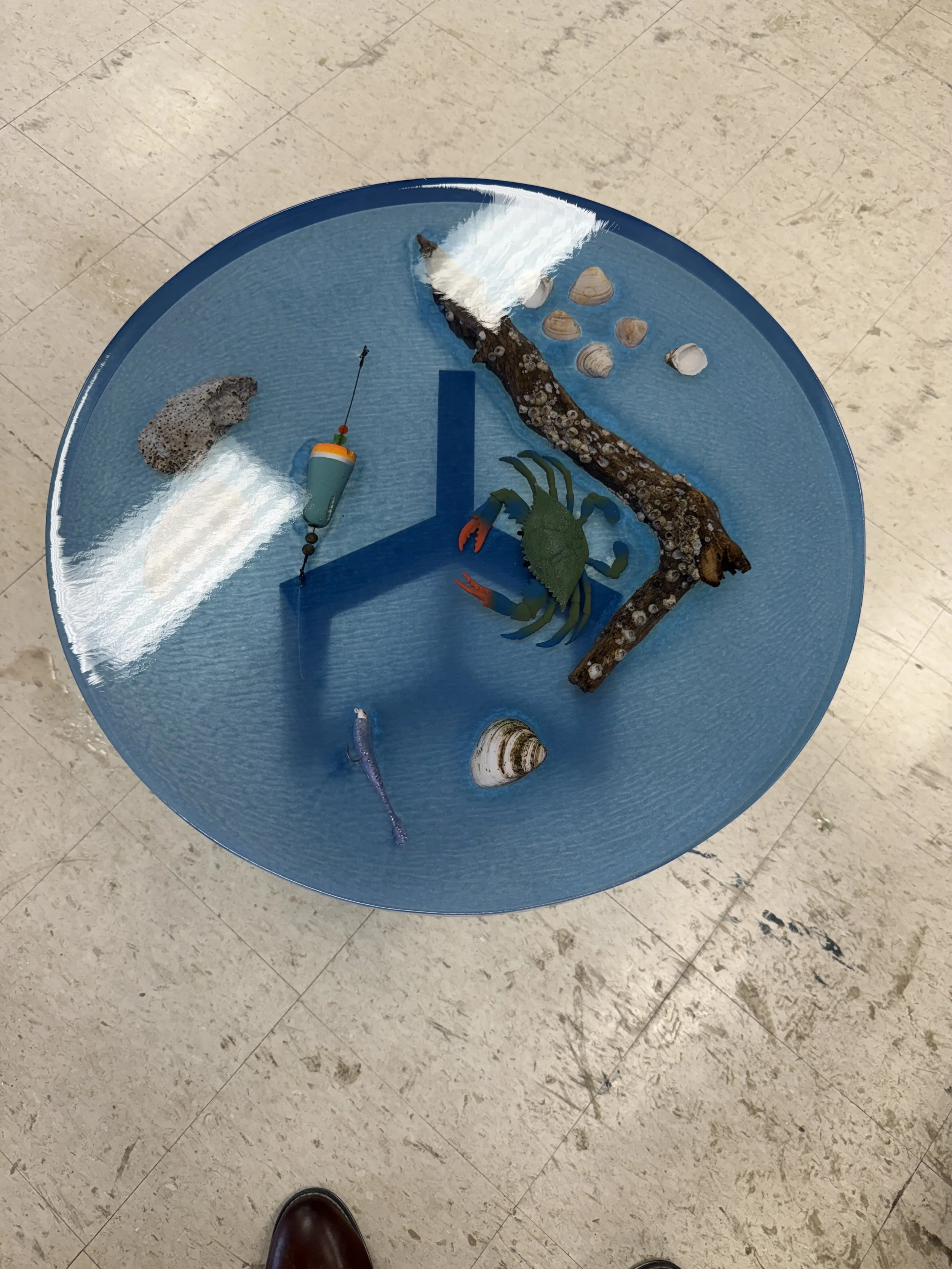 A round glass table with various sea life and fishing items submerged underneath, including a crab, a fish, seashells, and a piece of driftwood, seen from above.