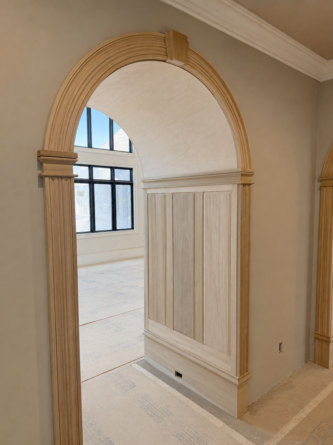 Interior view of a room with custom woodwork archway and wainscoting in progress, large window with black framing, and a beige wall.