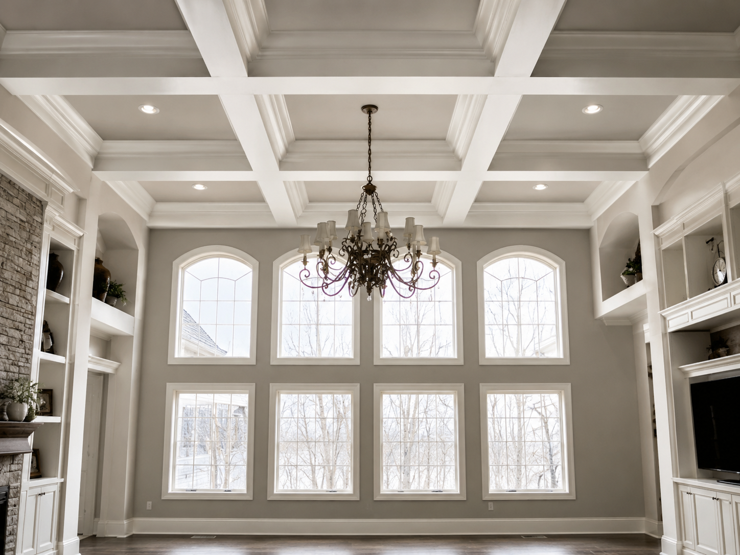 Empty living room with large grid of windows, white coffered ceiling, chandelier, built-in shelves with plants and decor, and a stone fireplace.