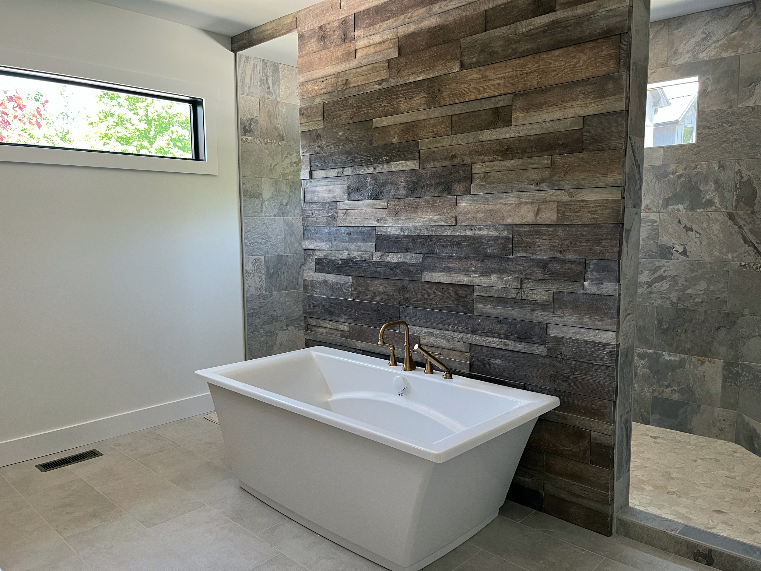 Bathroom with a white bathtub, a dark wooden accent wall, and gray tile walls with a window showing green foliage outside.