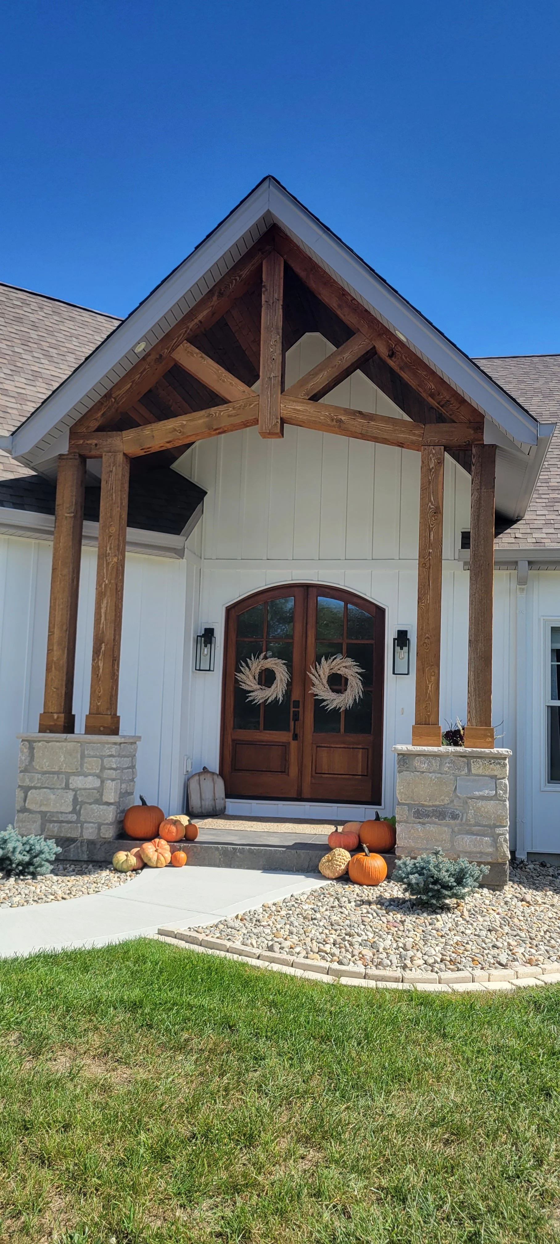 Front porch of a house decorated with pumpkins and gourds, with wreaths on the door, and a small garden bed with low shrubs and pebble mulch.