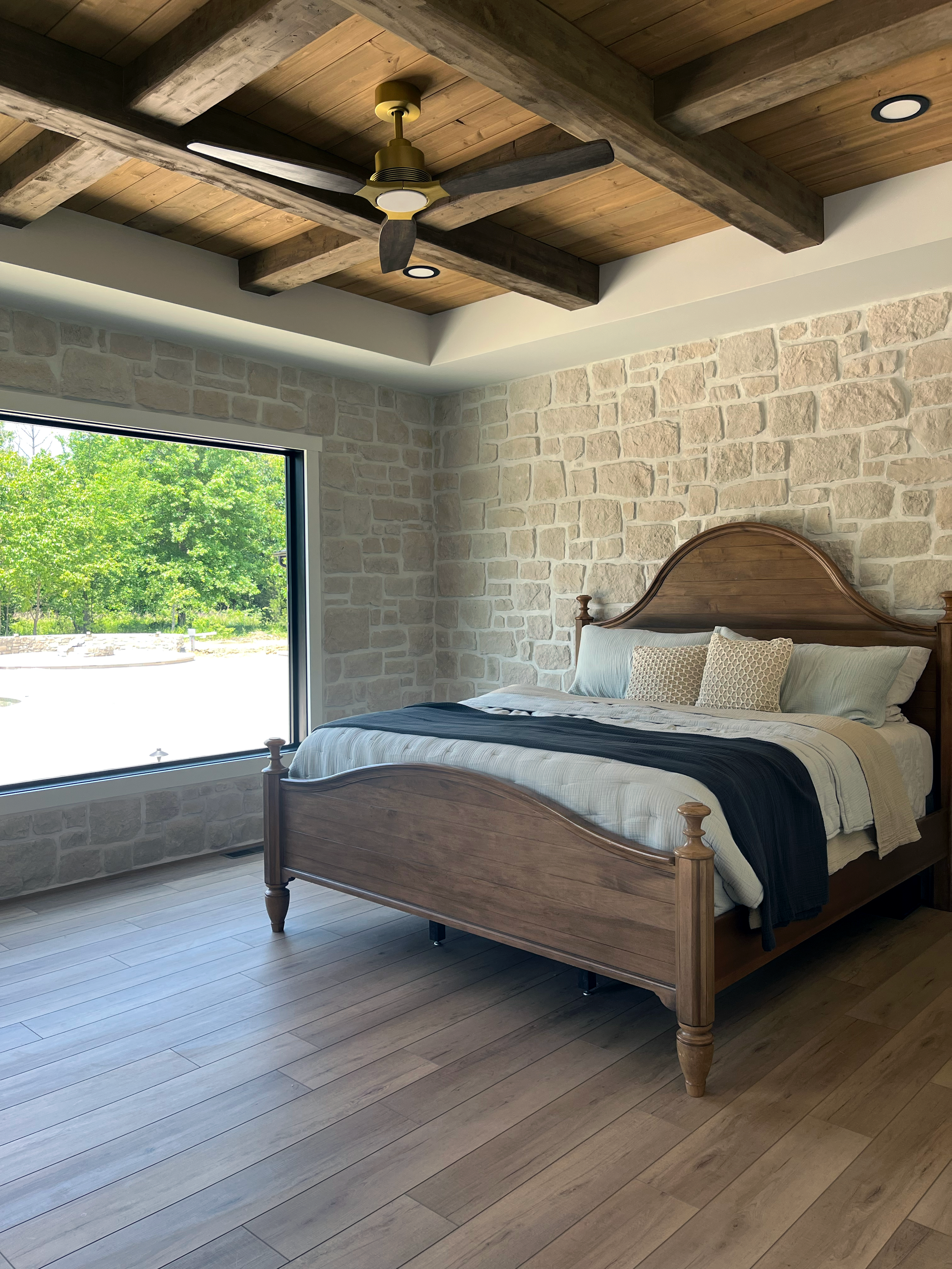 A bedroom with a wooden bed, pillows, a window with a view of green trees, and a ceiling with exposed wooden beams and a ceiling fan.