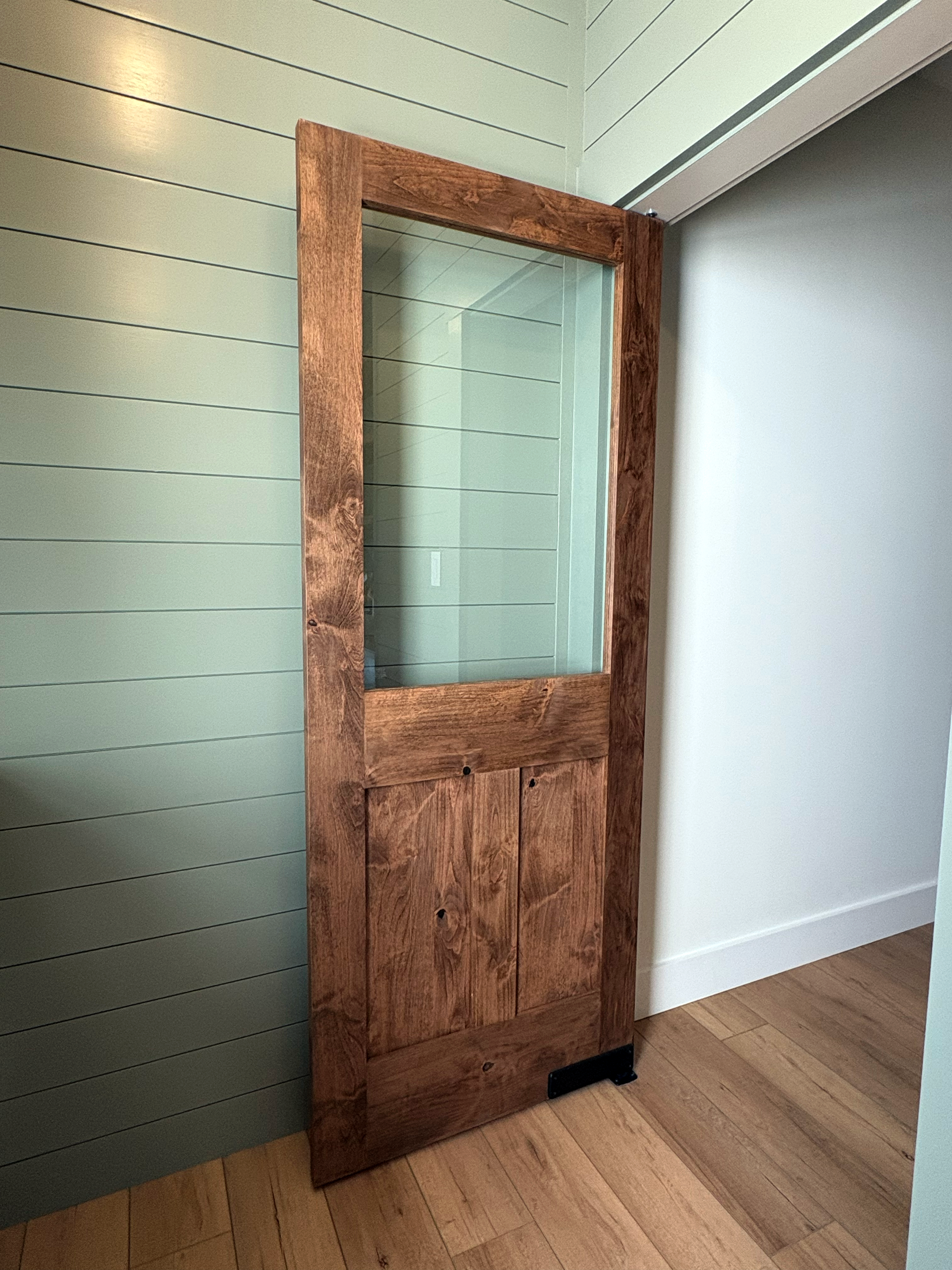 A wooden display case with a glass front, standing against a light green wall with horizontal paneling, with hardwood flooring underneath.