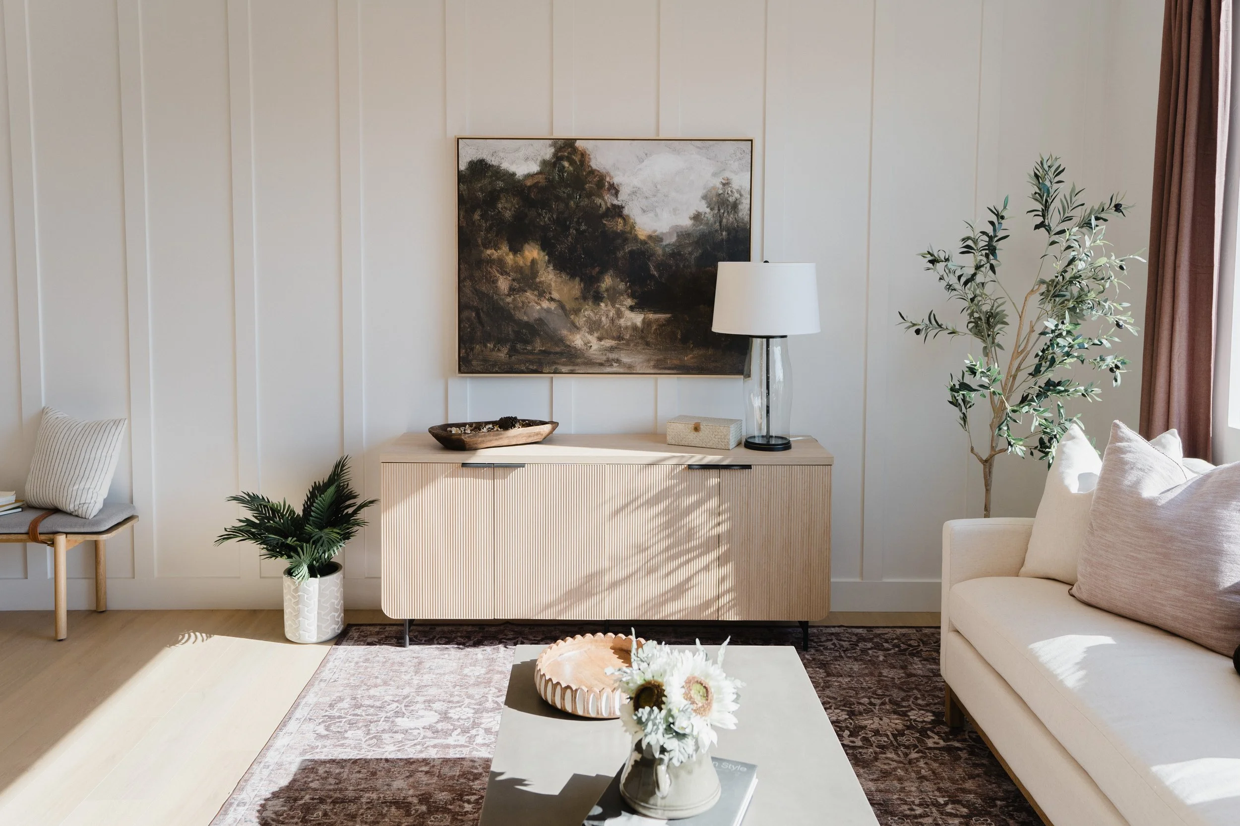 Modern living room with a beige sofa, potted plants, a sideboard with a lamp, artwork, and a coffee table with a vase of flowers.