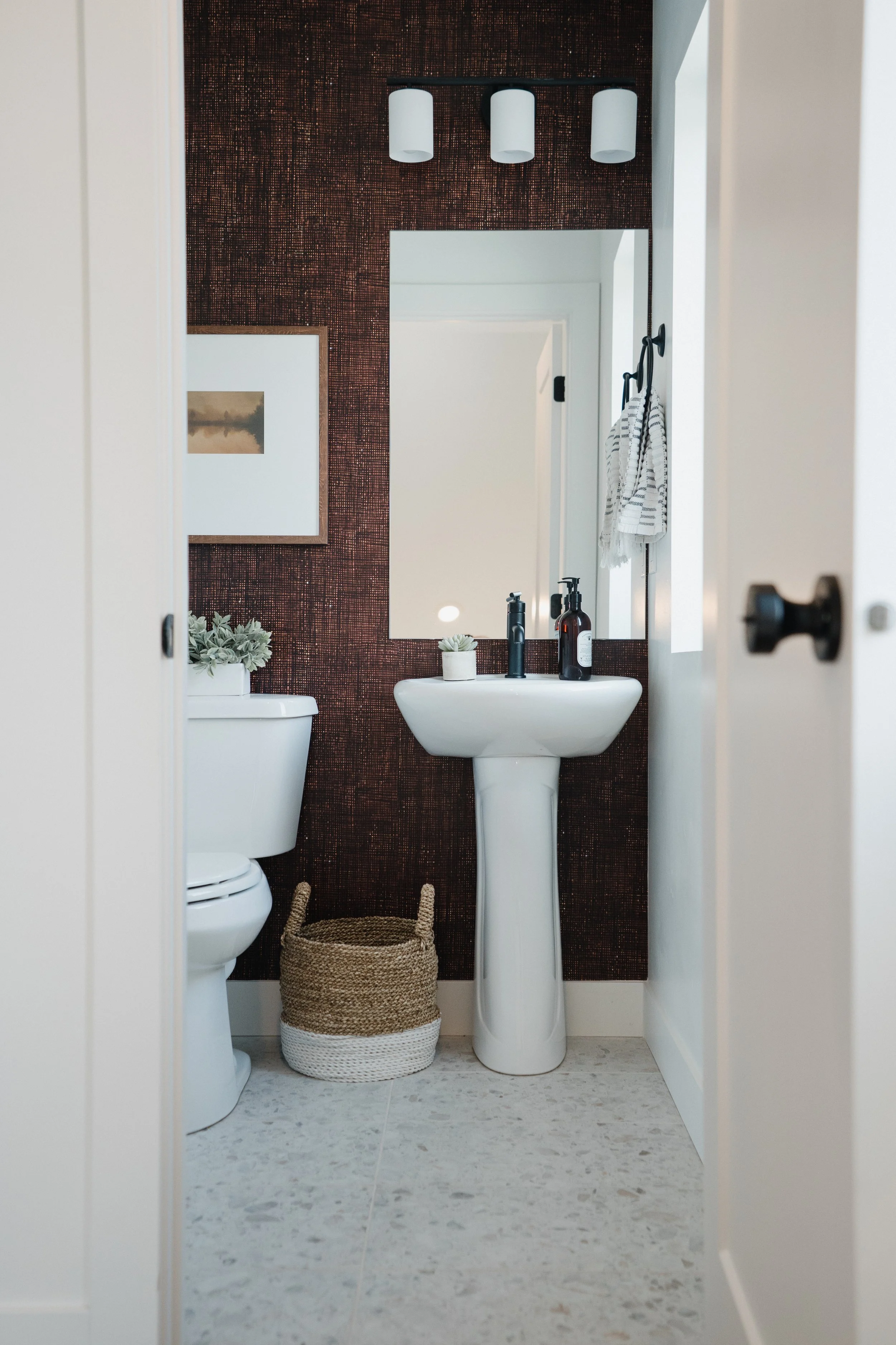A small bathroom with a white toilet and pedestal sink, a mirror, a brown textured accent wall, a small potted plant on the sink, a basket on the floor, a framed picture on the wall, and a window with a striped hand towel hanging on a hook.