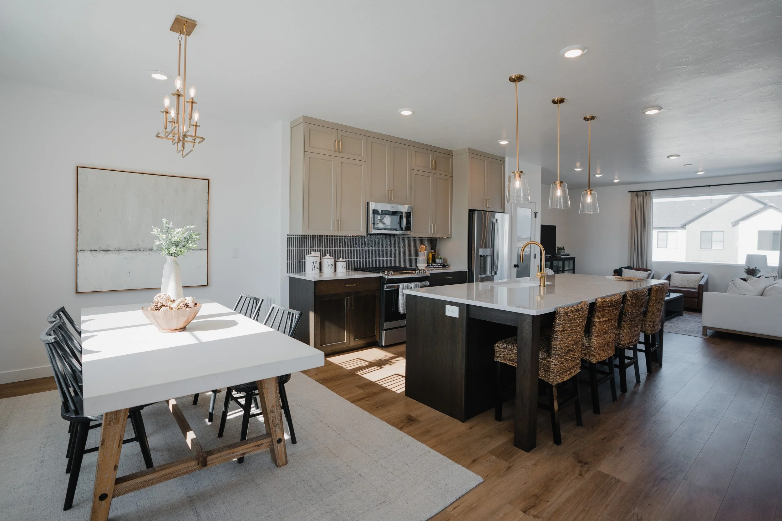 Open-concept kitchen and dining area with white walls, wooden flooring, beige cabinets, stainless steel appliances, a large kitchen island with wicker chairs, and a dining table with black chairs. Sunlight coming through large windows illuminating th