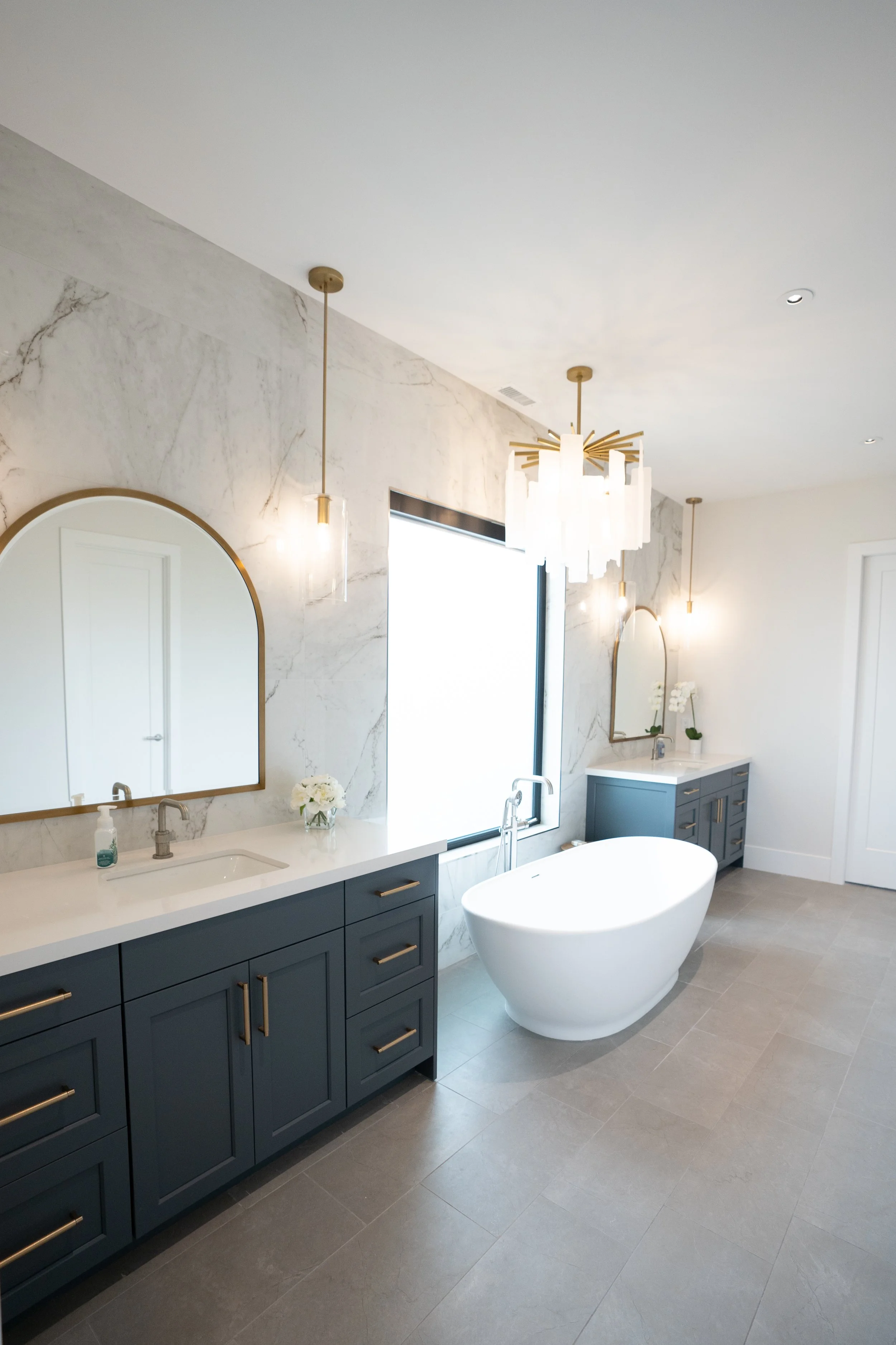Modern bathroom with blue vanities, white countertops, oval bathtub, marble wall, and gold fixtures.