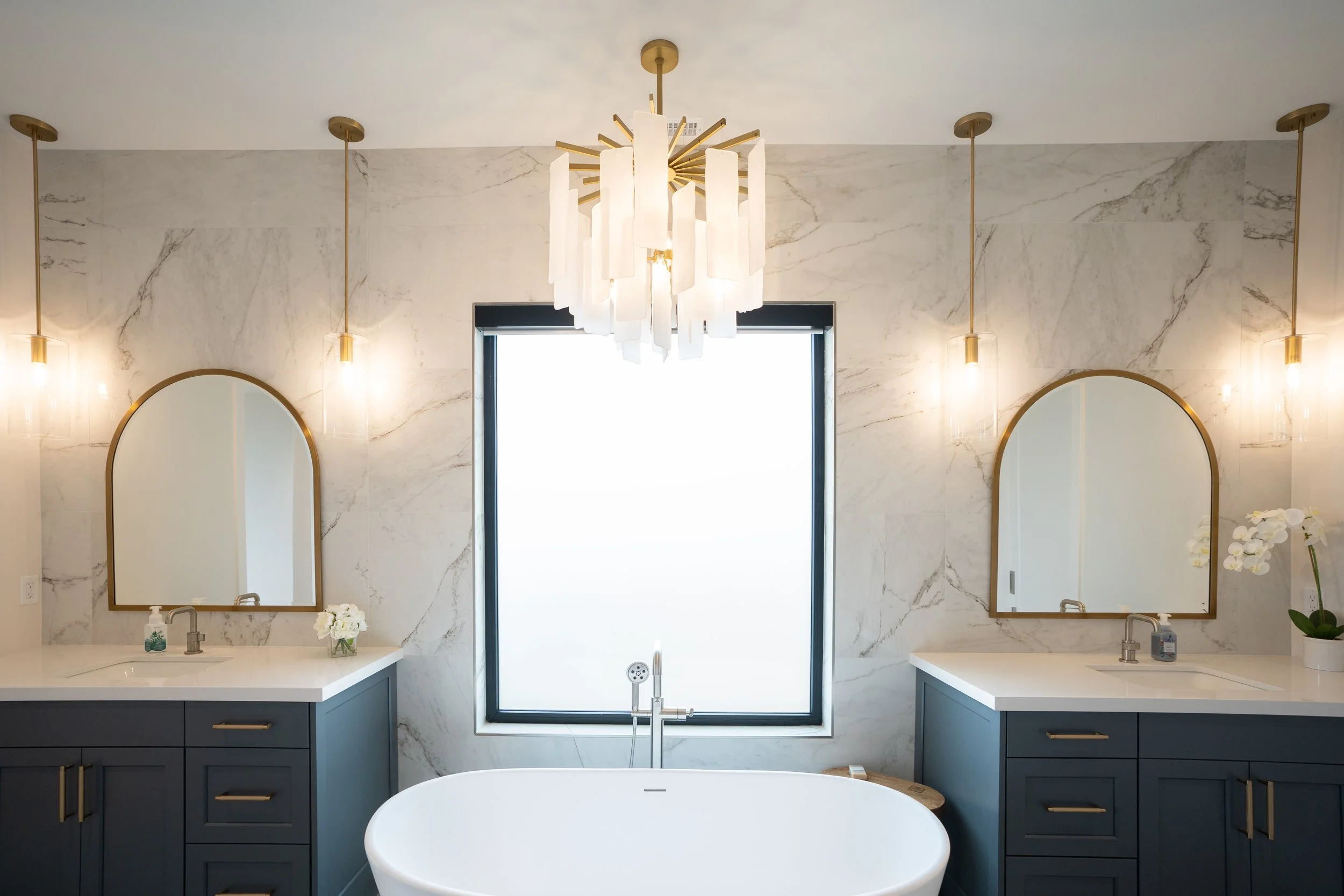 Modern bathroom with double sinks, dark blue cabinets, white marble walls, a central bathtub, and a large window in the background.