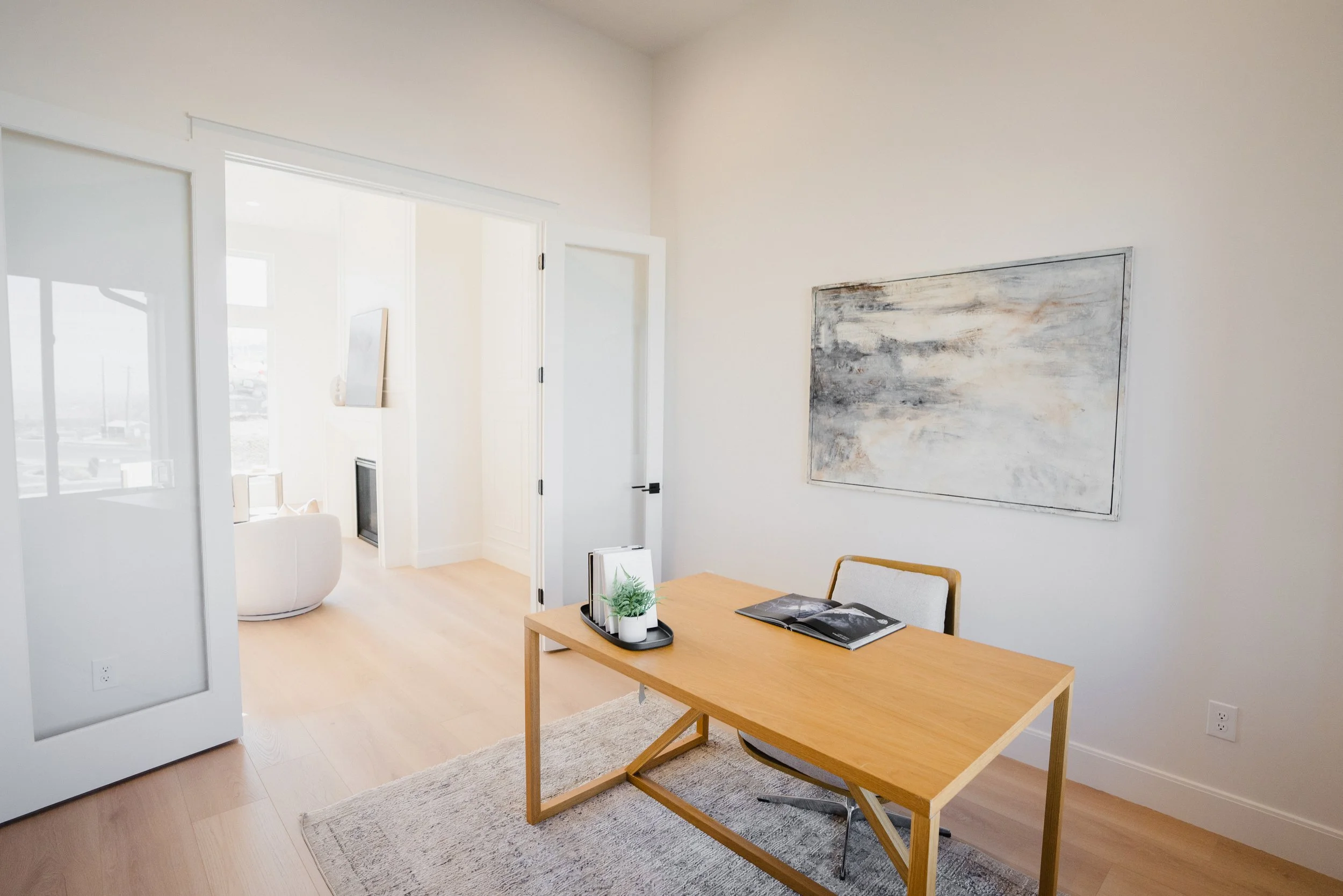 Minimalist office with a wooden desk, a chair, a large painting on the wall, and a view into a bright living room with a fireplace and modern decor.