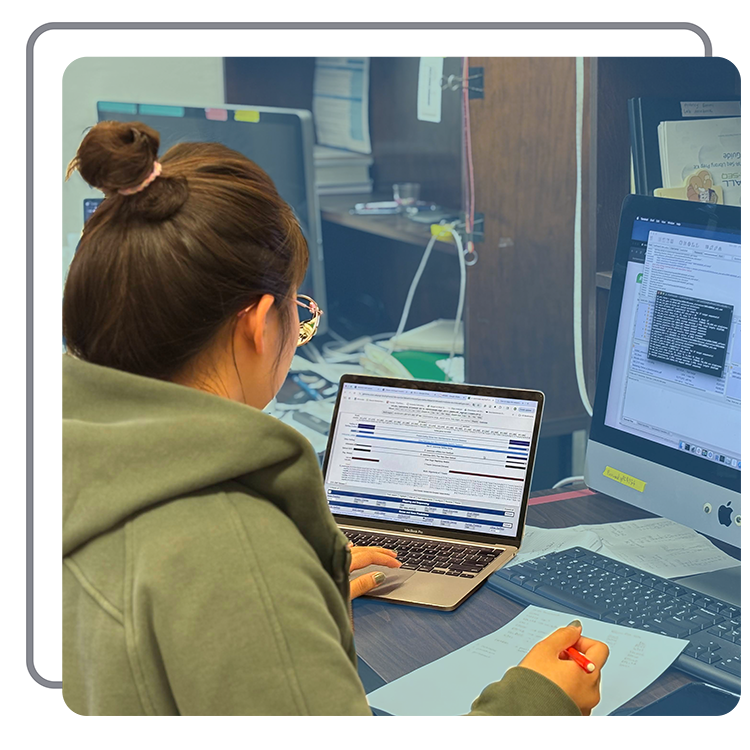 A woman with brown hair in a bun, wearing glasses and a green hoodie, working at a desk with a laptop, a desktop computer, and papers, in an office environment.