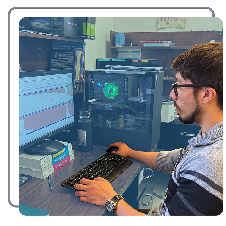 A man with glasses and a gray striped sweater working at a desk with dual monitors, a computer tower with RGB lighting, a water bottle, pen, and stacked books in a cluttered office with shelves and posters.