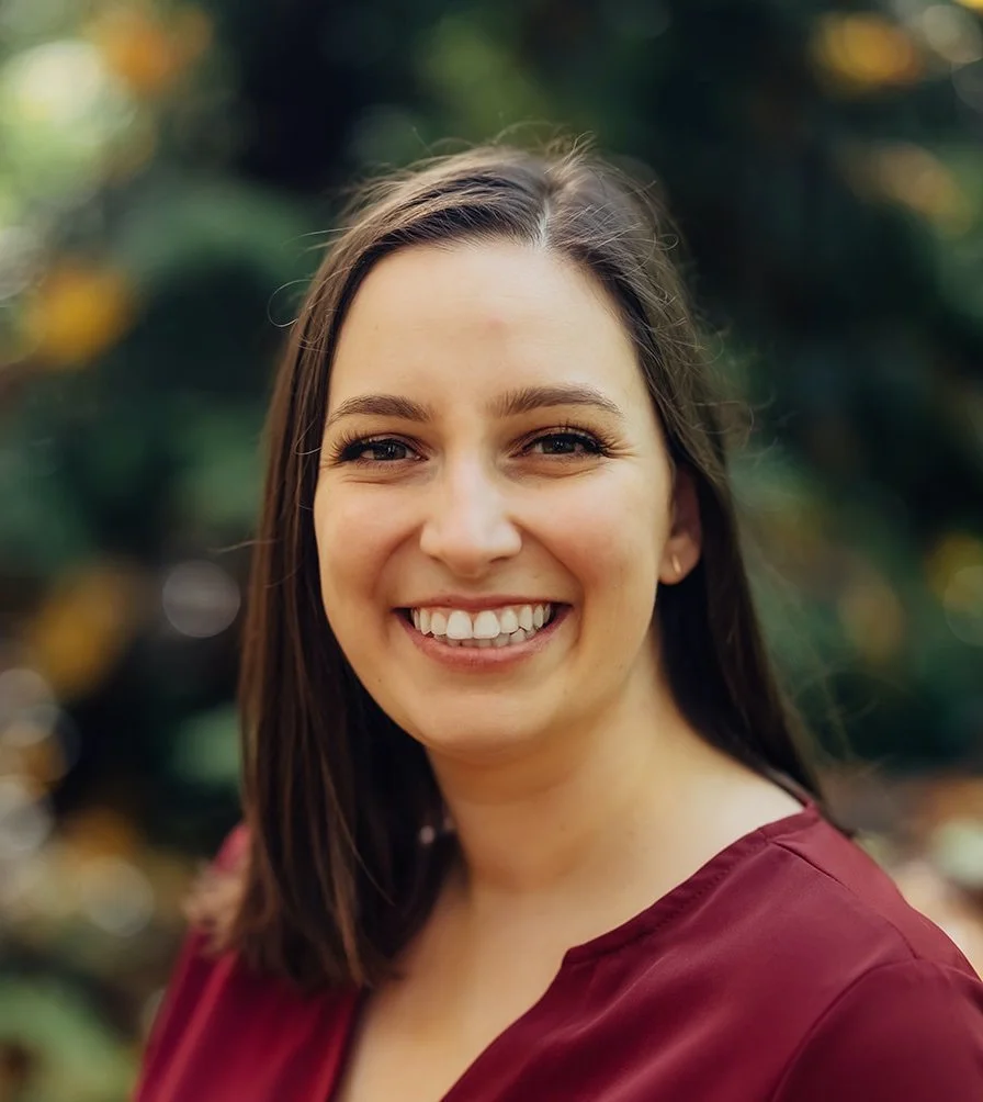 A woman smiling outdoors with blurred greenery in the background.