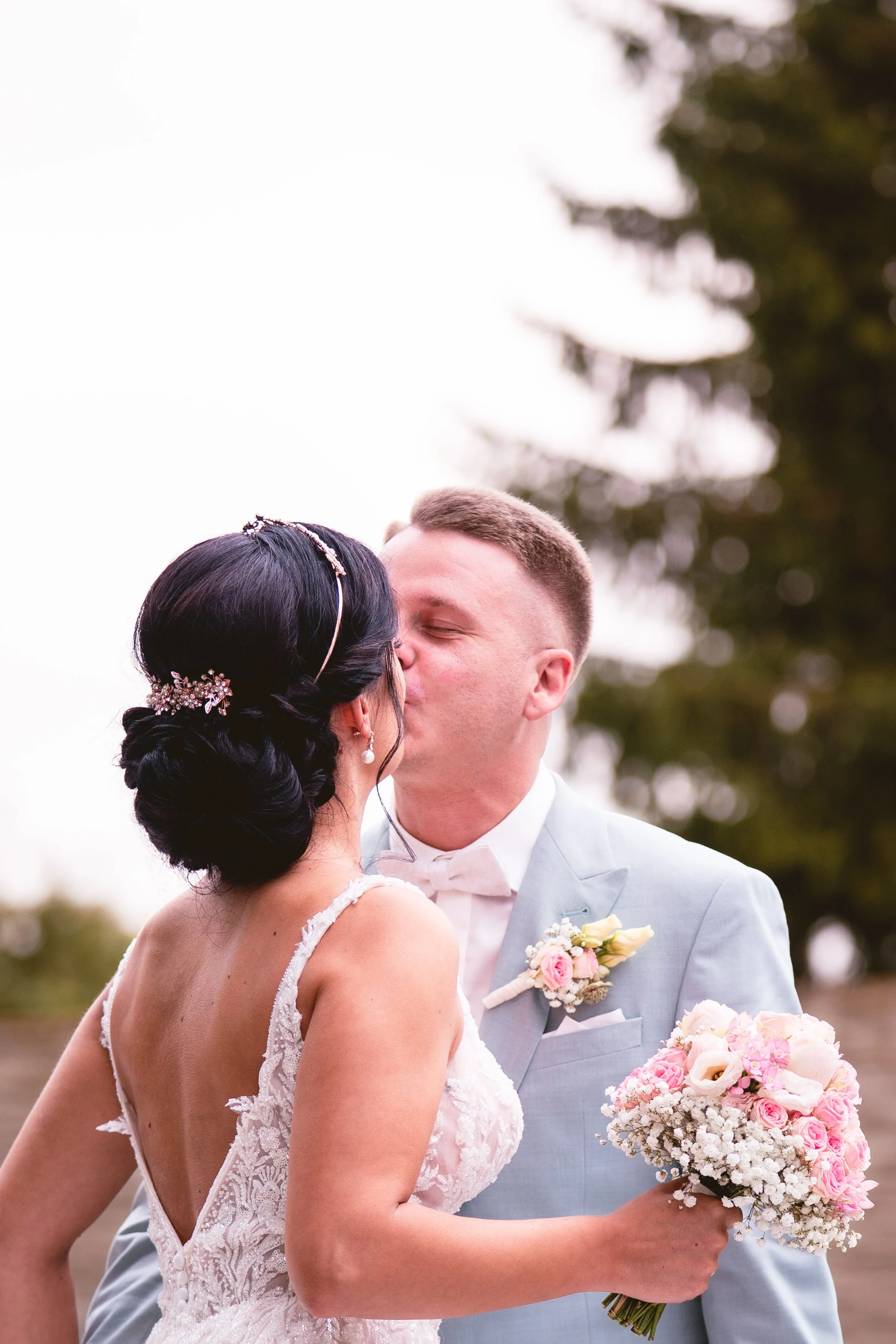 Ein Brautpaar bei ihrer Hochzeit, die Braut hält einen Blumenstrauß, der Bräutigam trägt einen Anzug mit Fliege, im Hintergrund sind Bäume zu sehen.