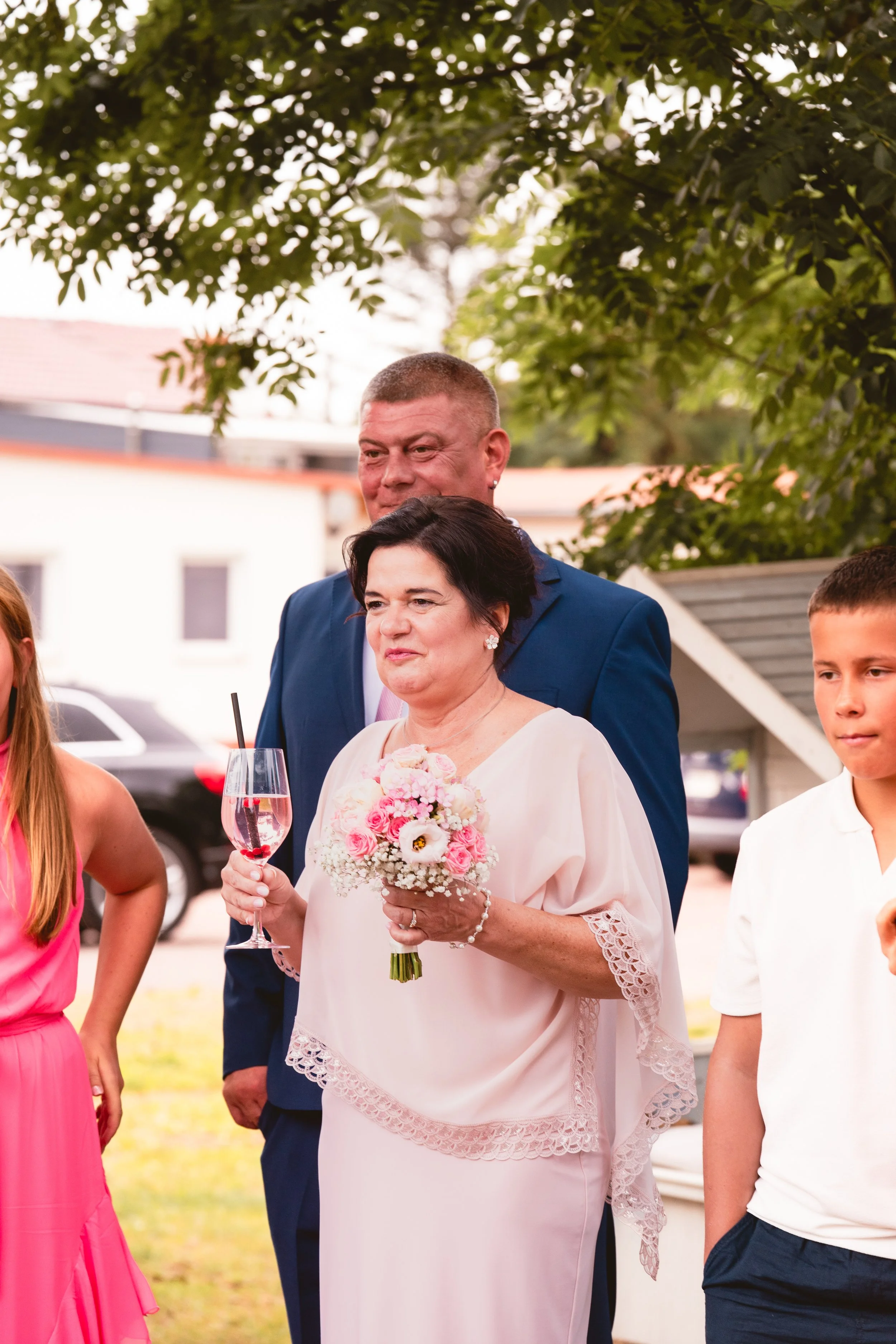 Eine Gruppe von Menschen bei einer Feier im Freien, im Mittelpunkt eine Frau in einem weißen Kleid mit Spitze, die ein Blumenbouquet und ein Glas mit rosa Getränk hält, dahinter ein Mann in blauem Anzug, umgeben von weiteren Personen.
