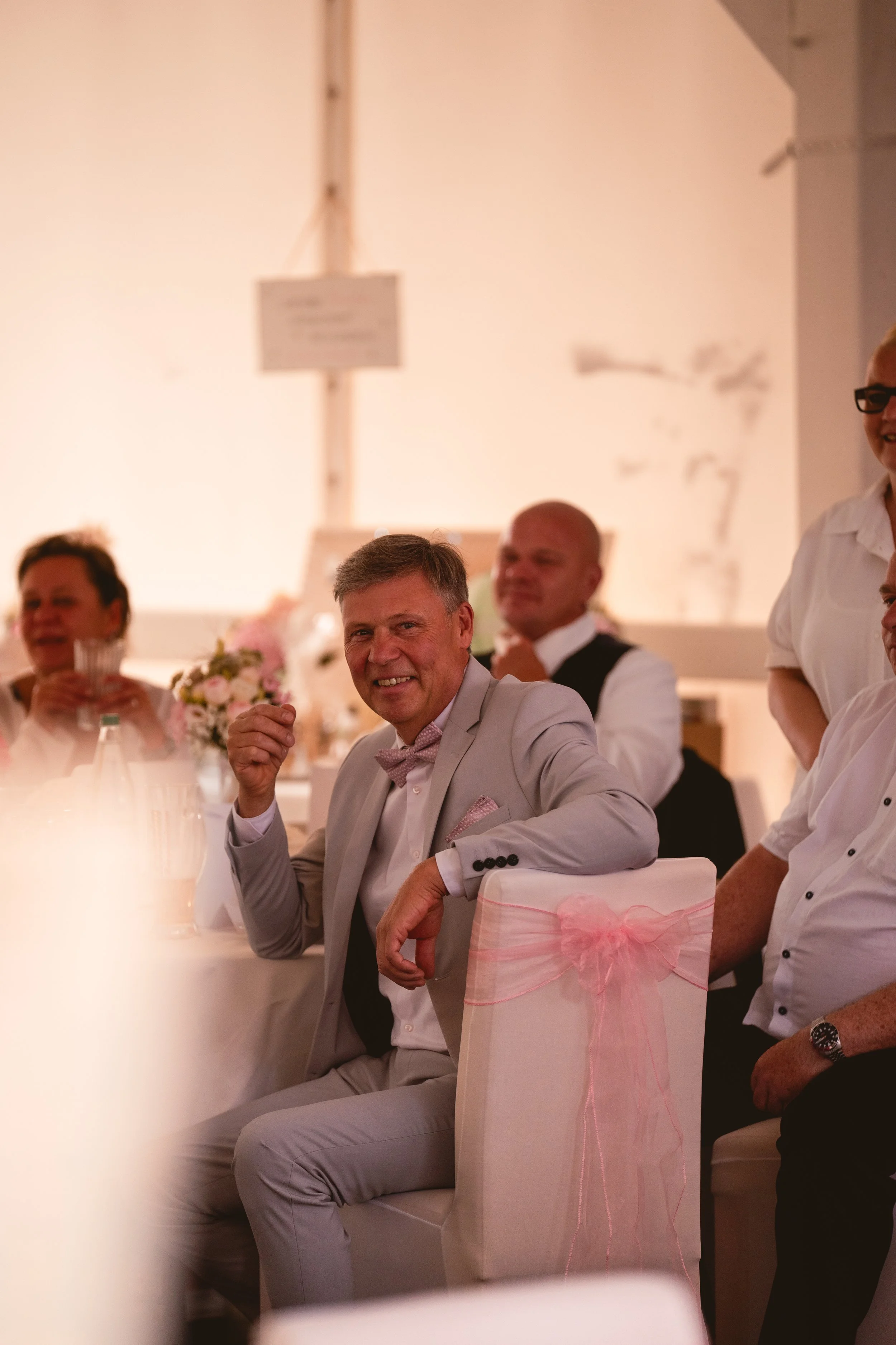 Menschen sitzen an einem festlich gedeckten Tisch bei einer Veranstaltung, der Mann im Vordergrund trägt einen hellgrauen Anzug und ein lila Hemd, er lächelt und schaut in die Kamera, während andere Personen im Hintergrund sich unterhalten und Geträn