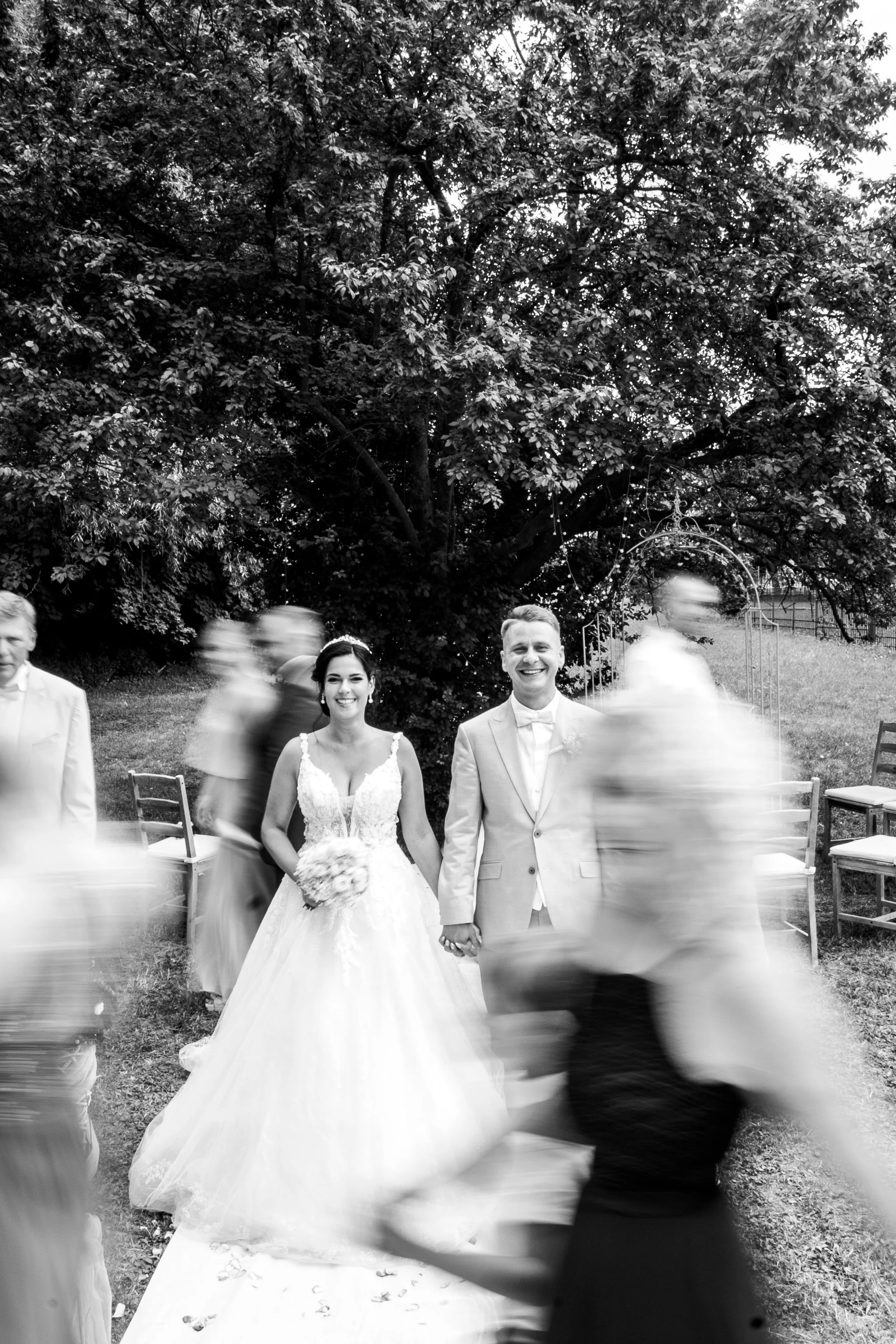 Brautpaar mit Trauzeugen bei einer Hochzeit im Freien, das Bild ist verschwommen, während Gäste vorbeigehen.