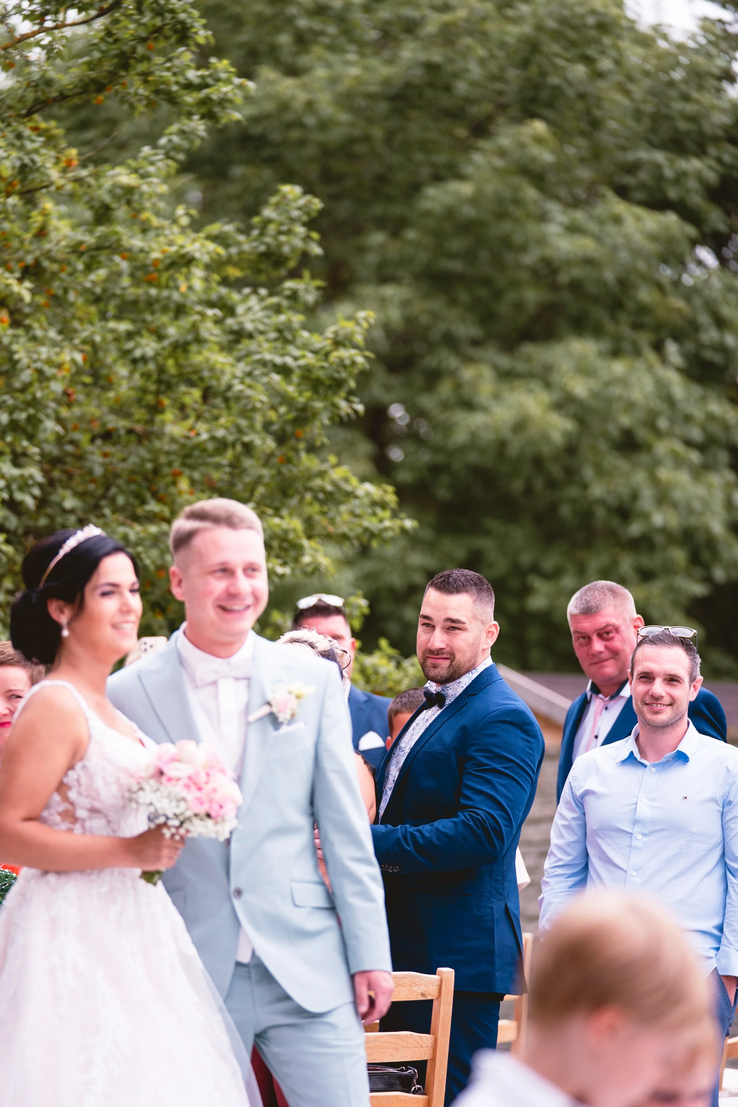 Eine Gruppe von Menschen bei einer Hochzeit im Freien, die Braut in einem weißen Kleid mit Blumenstrauß, die Männer in Anzügen, der Hintergrund mit Bäumen und blauer Himmel.