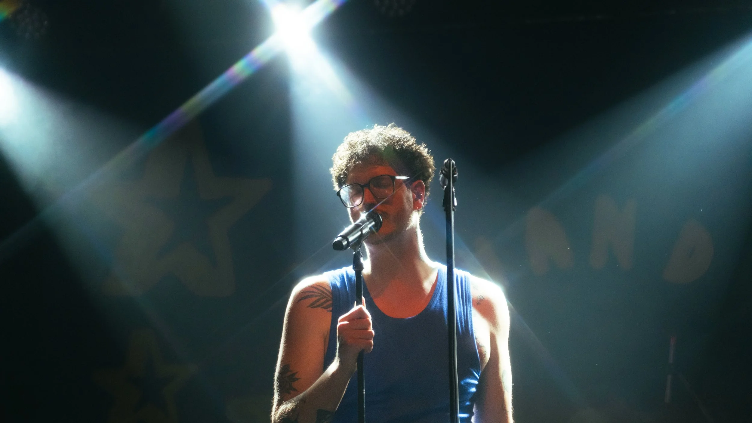Ein Musiker mit Brille, Tattoos und einem blauen Tanktop singt in einem dunklen Raum unter starken Lichtstrahlen, die von oben auf ihn gerichtet sind.