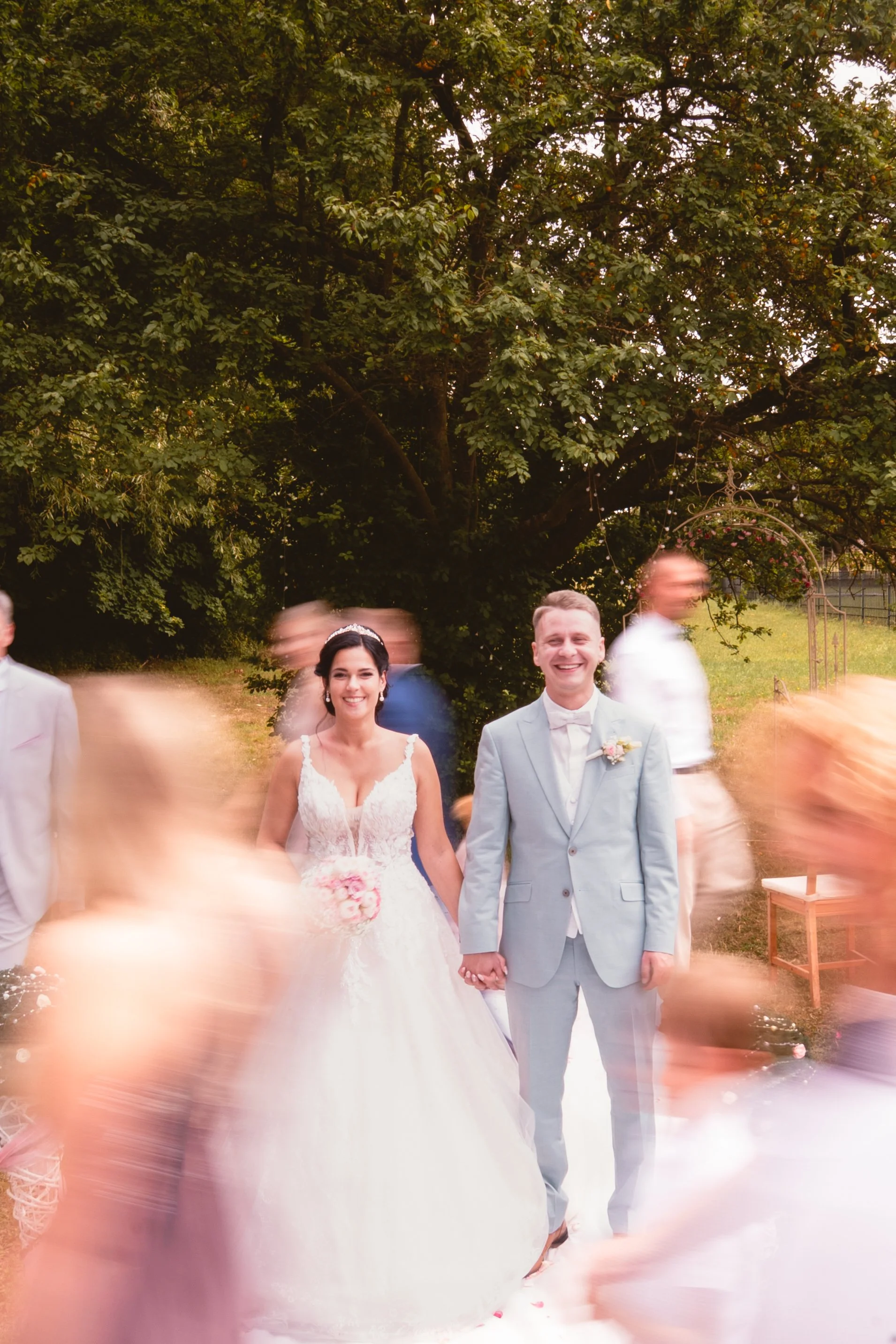 Ein Brautpaar, ein Mann in einem hellblauen Anzug und eine Frau in einem weißen Hochzeitskleid, steht Hand in Hand im Freien unter einem Baum bei Sonnenuntergang. Es herrscht Bewegung um sie herum, vermutlich Gäste bei einer Hochzeit.