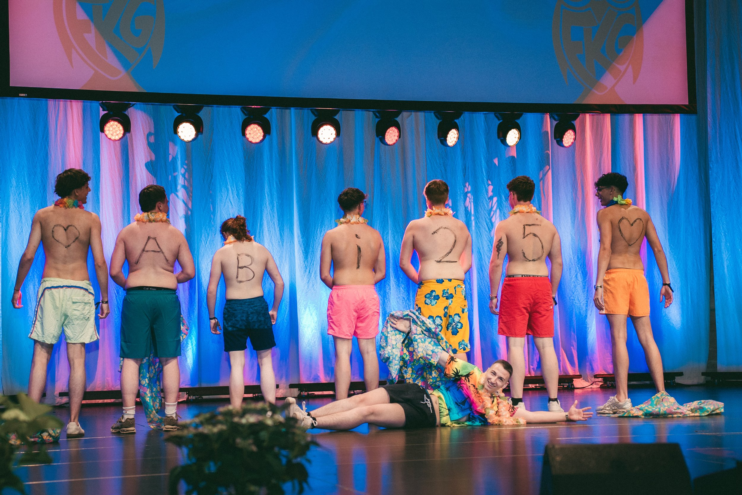 Gruppe junger Menschen in bunten Shorts und Blumenketten auf einer Bühne, some mit aufgemalten Buchstaben und Herzen auf dem Rücken, ein Mann liegt am Boden mit Arm ausgestreckt, im Hintergrund eine große Leinwand mit einem Logo. Es scheint eine inszenierte Show oder Performance zu sein.