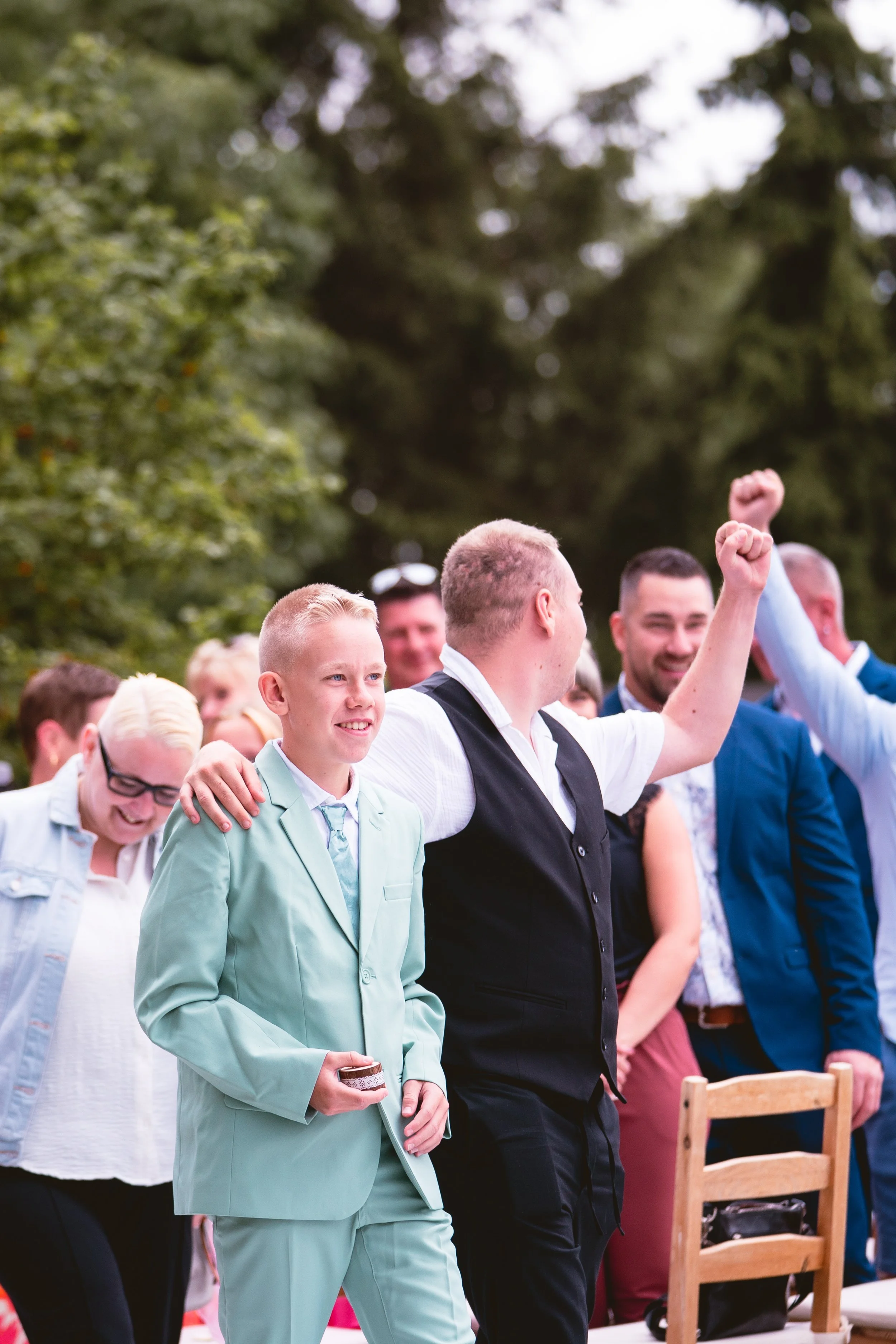 Gruppe von jungen Menschen beim Fest, einige heben die Fäuste in die Luft, im Hintergrund sind Bäume und ein Himmel sichtbar.