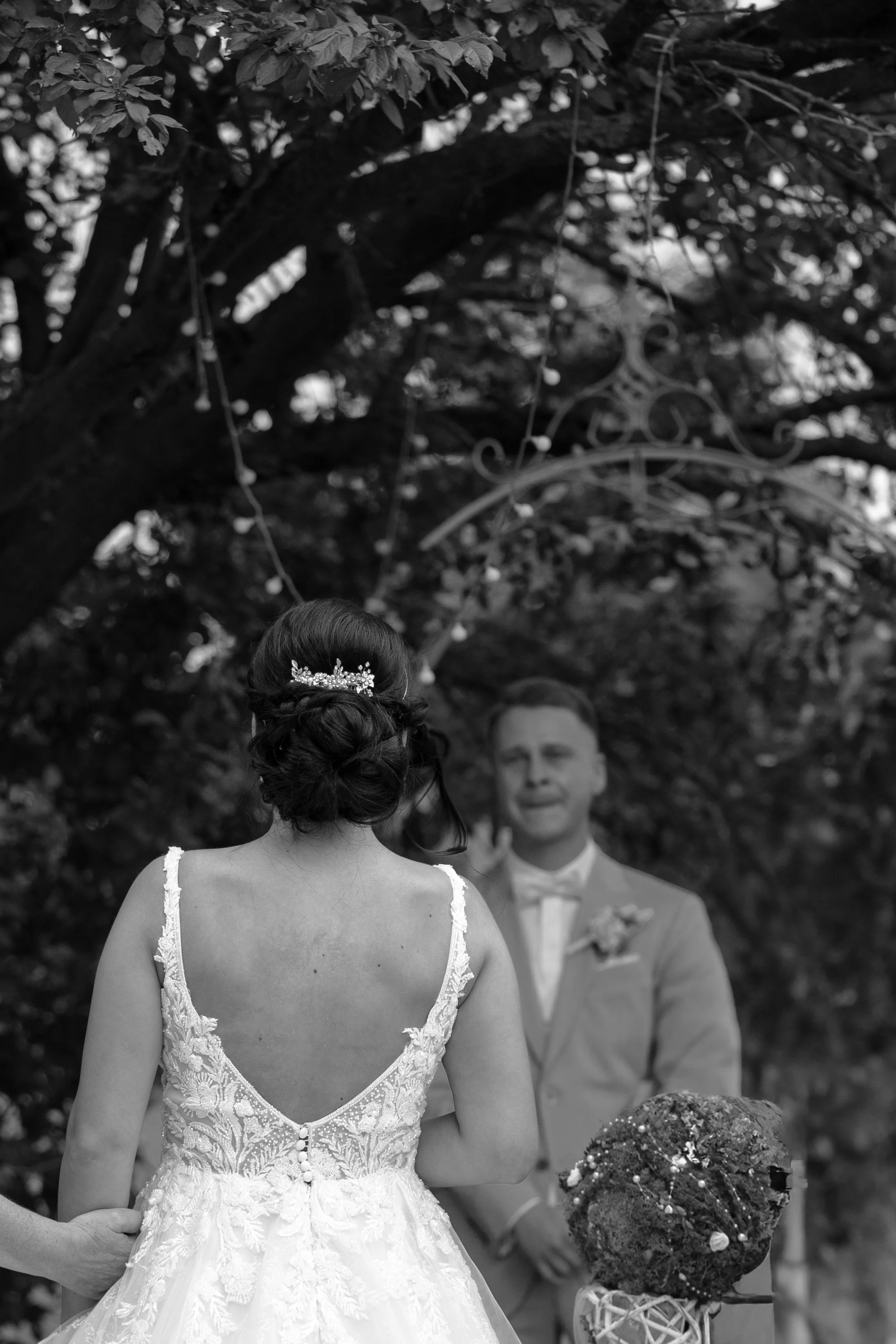Braut in einem weißen Spitzenkleid bei einer Hochzeit, im Hintergrund ein Bräutigam in einem Anzug, umgeben von Bäumen und Dekorationen.