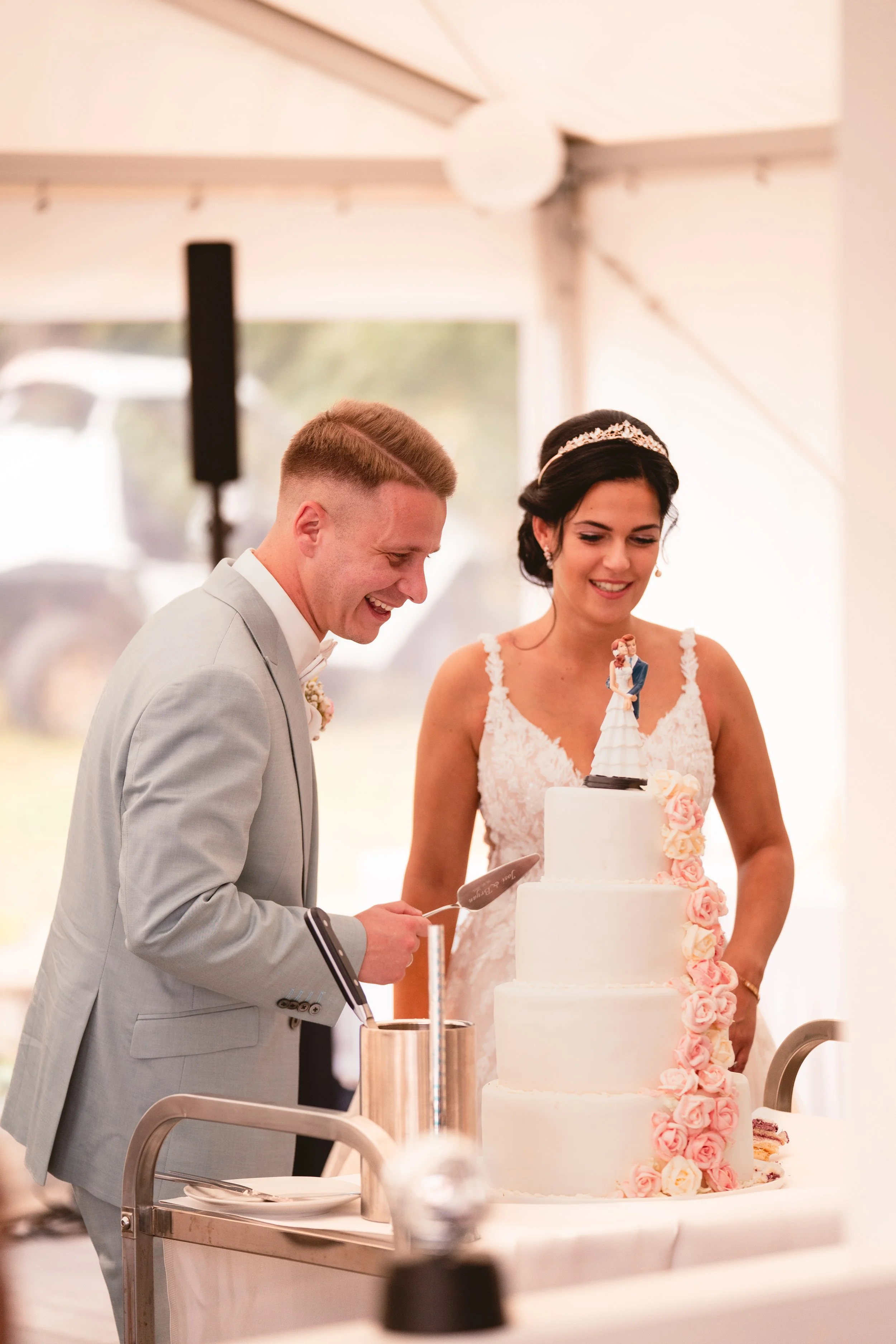 Ein Brautpaar beim Anschneiden einer Hochzeitstorte in einem festlich dekorierten Raum, der Blick auf die Hochzeittorte mit Rosenverzierung.
