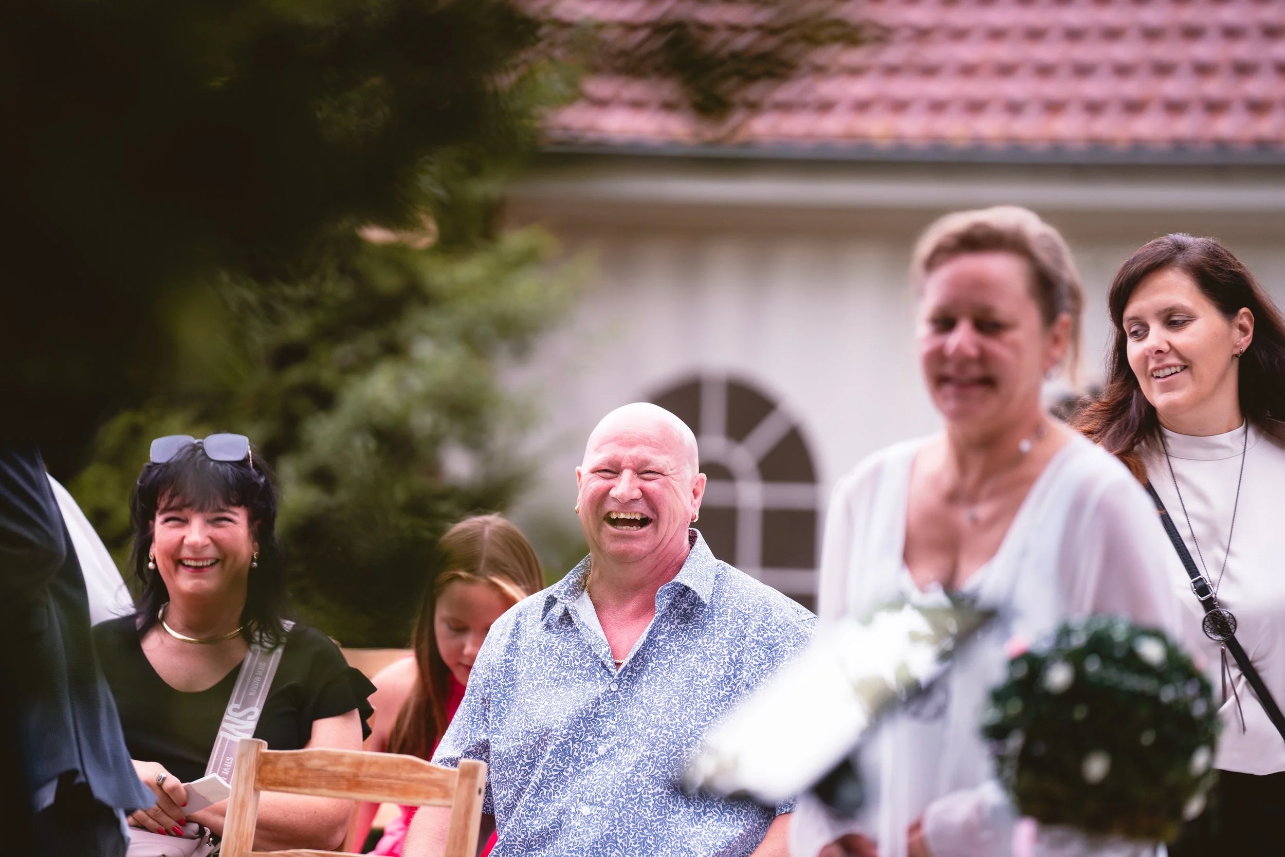 Gruppe lachender Menschen bei einem Treffen im Garten vor einem Haus mit rotem Dach, einige tragen Brillen und haben unterschiedliche Haarfarben.