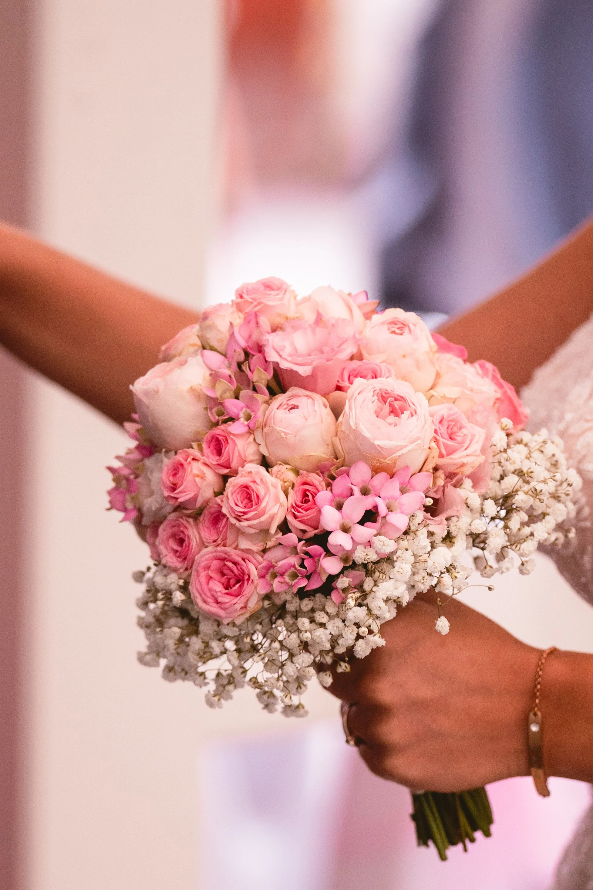 Ein Brautstrauß mit rosa und weißen Blumen wird gehalten, im Hintergrund ist eine Person sichtbar, die vermutlich eine Braut ist.
