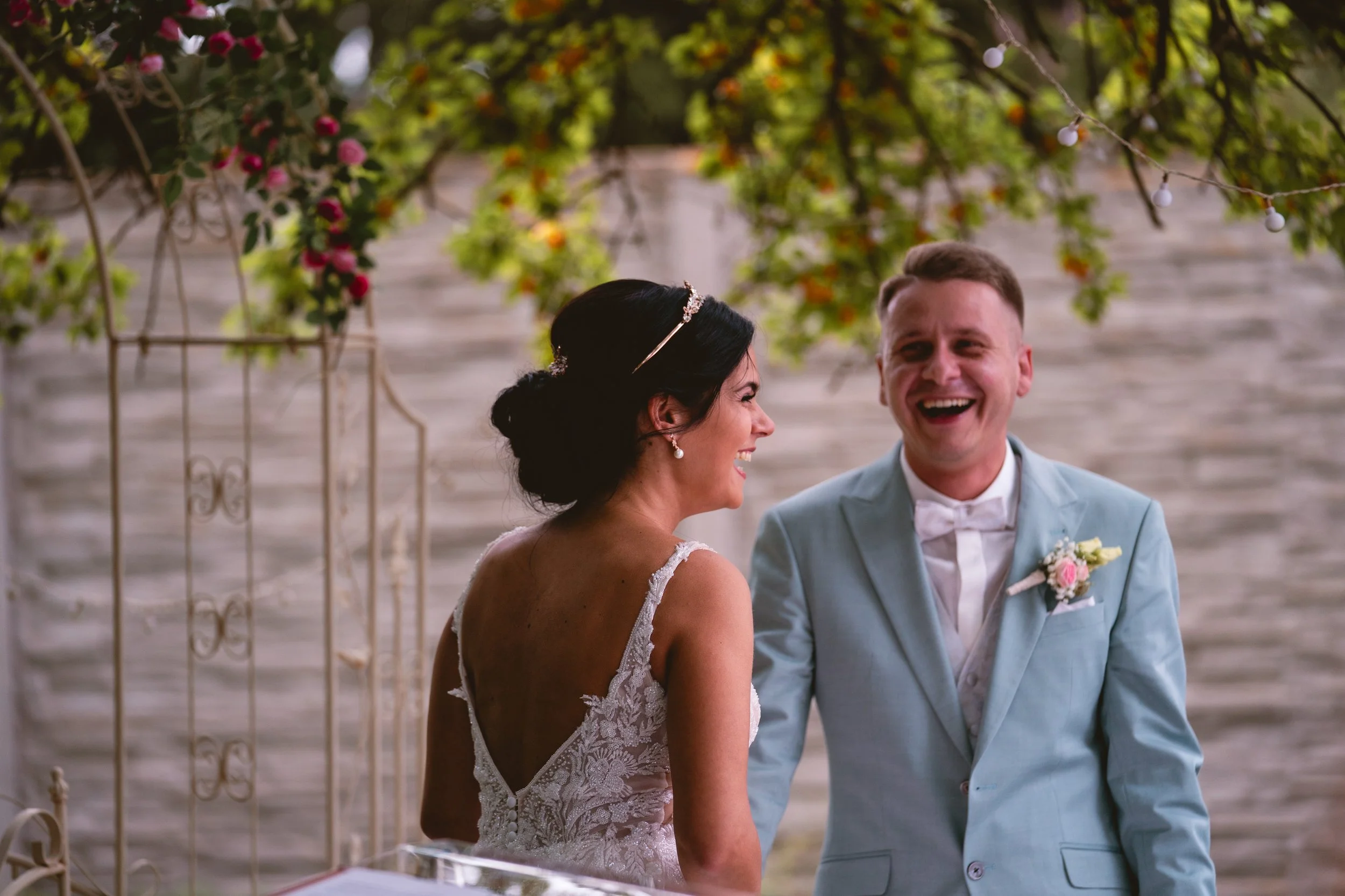 Ein Brautpaar bei einer Hochzeit, lachend, unter einem blühenden Baum im Freien.