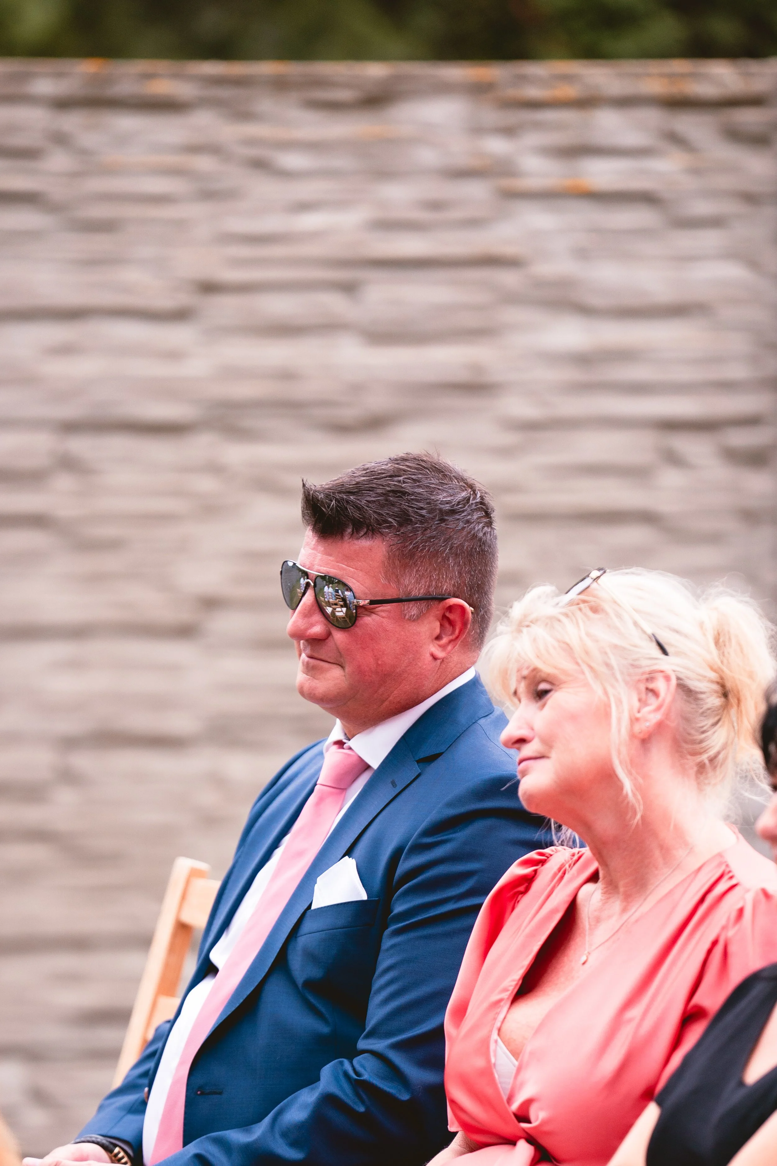 Ein Mann in einem blauen Anzug mit rosa Krawatte und Sonnenbrille sitzt neben einer Frau in einem roten Kleid bei einer Veranstaltung.