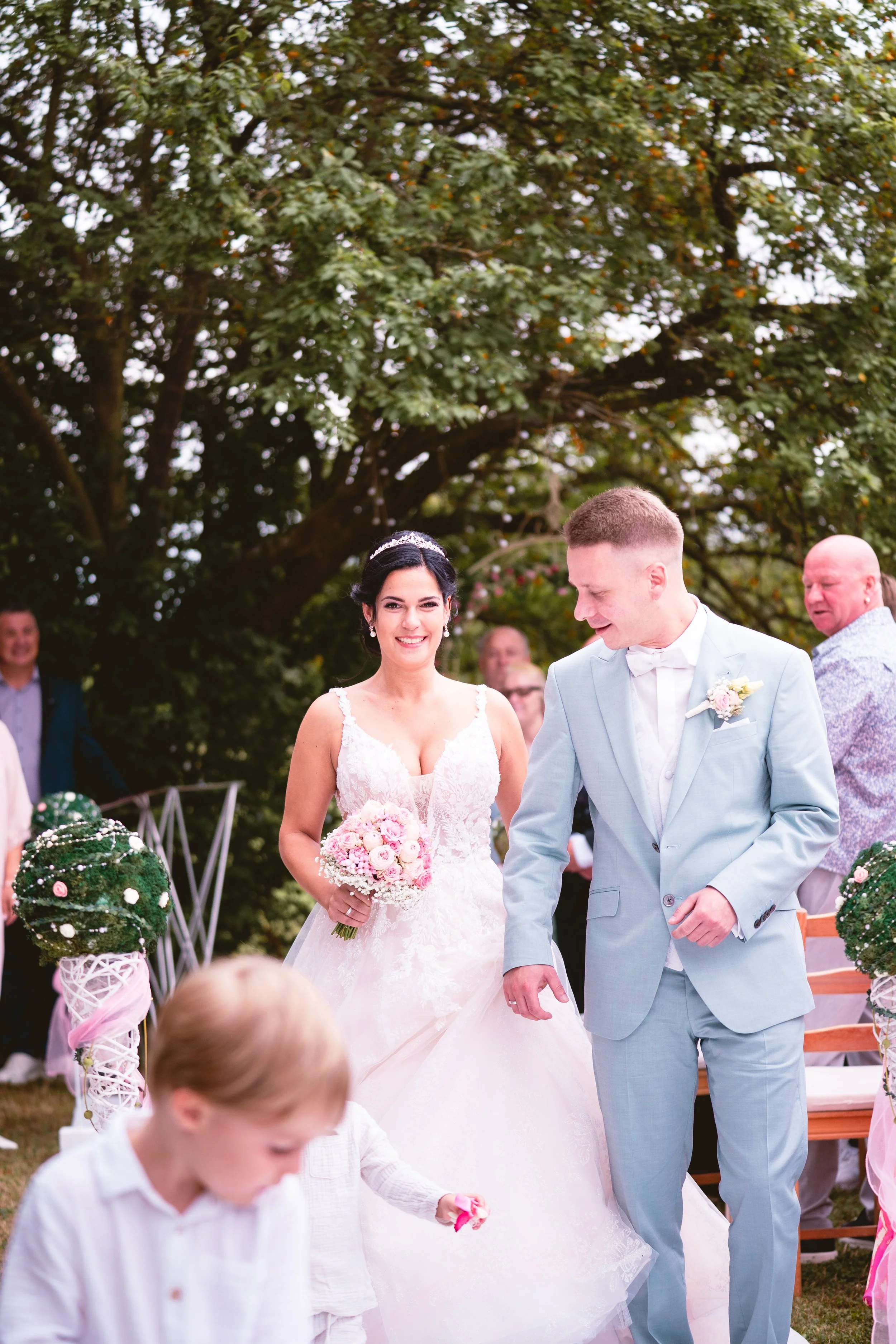 Braut und Bräutigam bei einer Hochzeit im Freien, mit Gästen und Dekorationen in Szenerie mit Bäumen im Hintergrund.