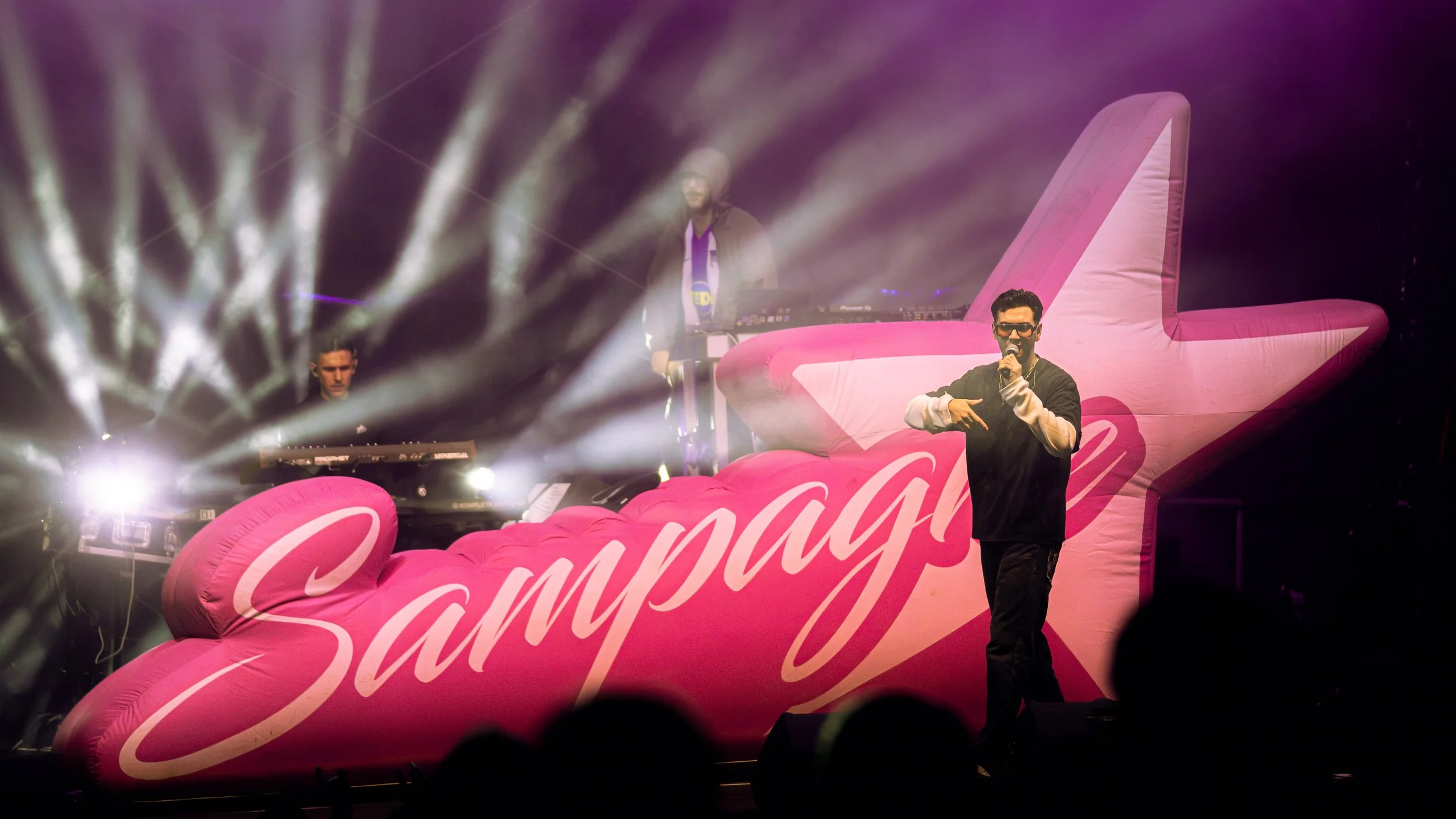 Ein Mann trägt Sonnenbrille und hält ein Mikrofon, während er auf einer pinkfarbenen, aufblasbaren Plattform mit dem Schriftzug 'CAMPAG' steht, mit einer DJ-Performance im Hintergrund und mehreren Scheinwerfern, die Lichtstrahlen projizieren.