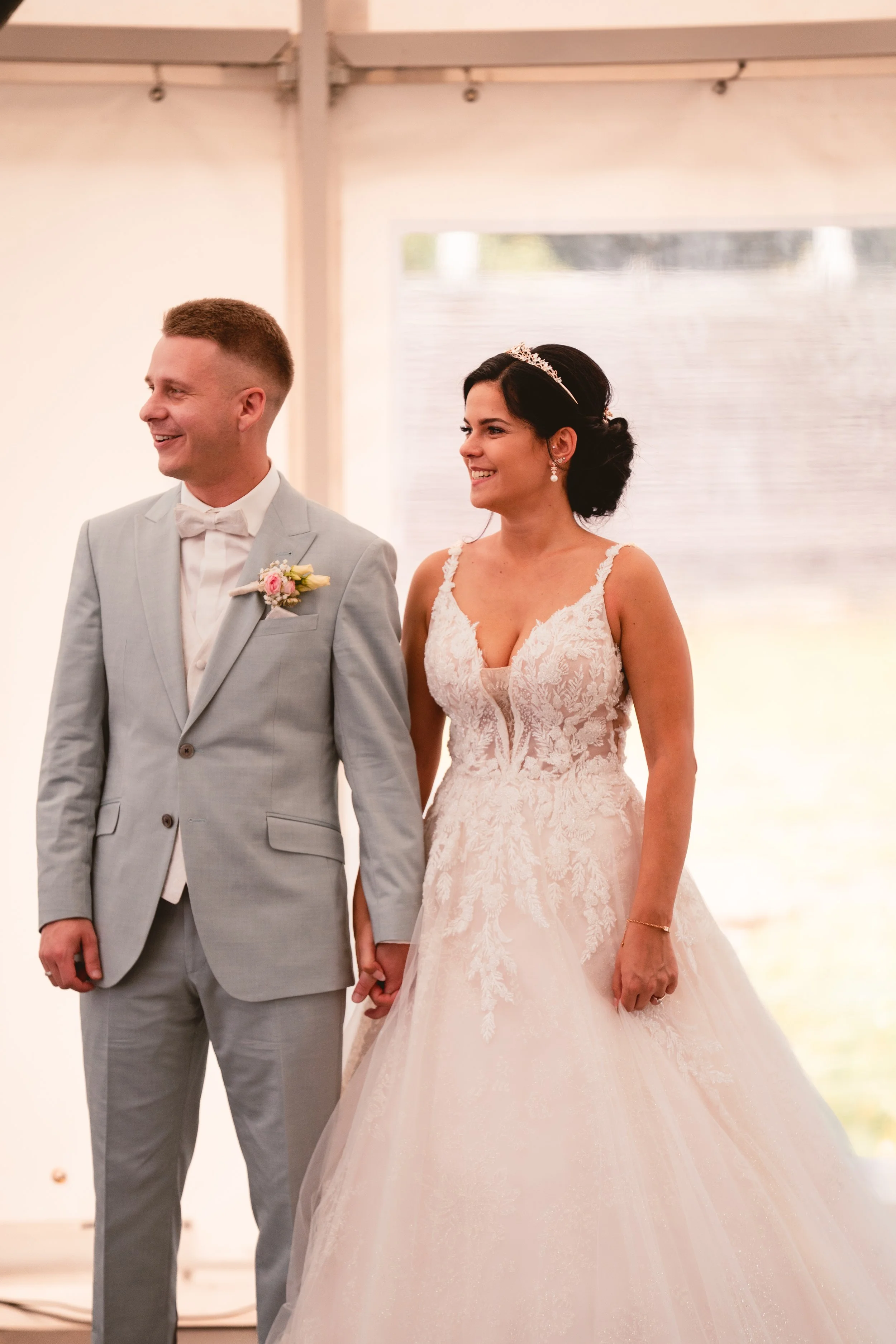 Ein Brautpaar bei einer Hochzeit, die Braut trägt ein weißes, Spitzenkleid mit Trägern und die Haare sind zu einem Dutt hochgesteckt, der Bräutigam trägt einen hellgrauen Anzug mit Fliege und einen Blumenstrauß am Revers.
