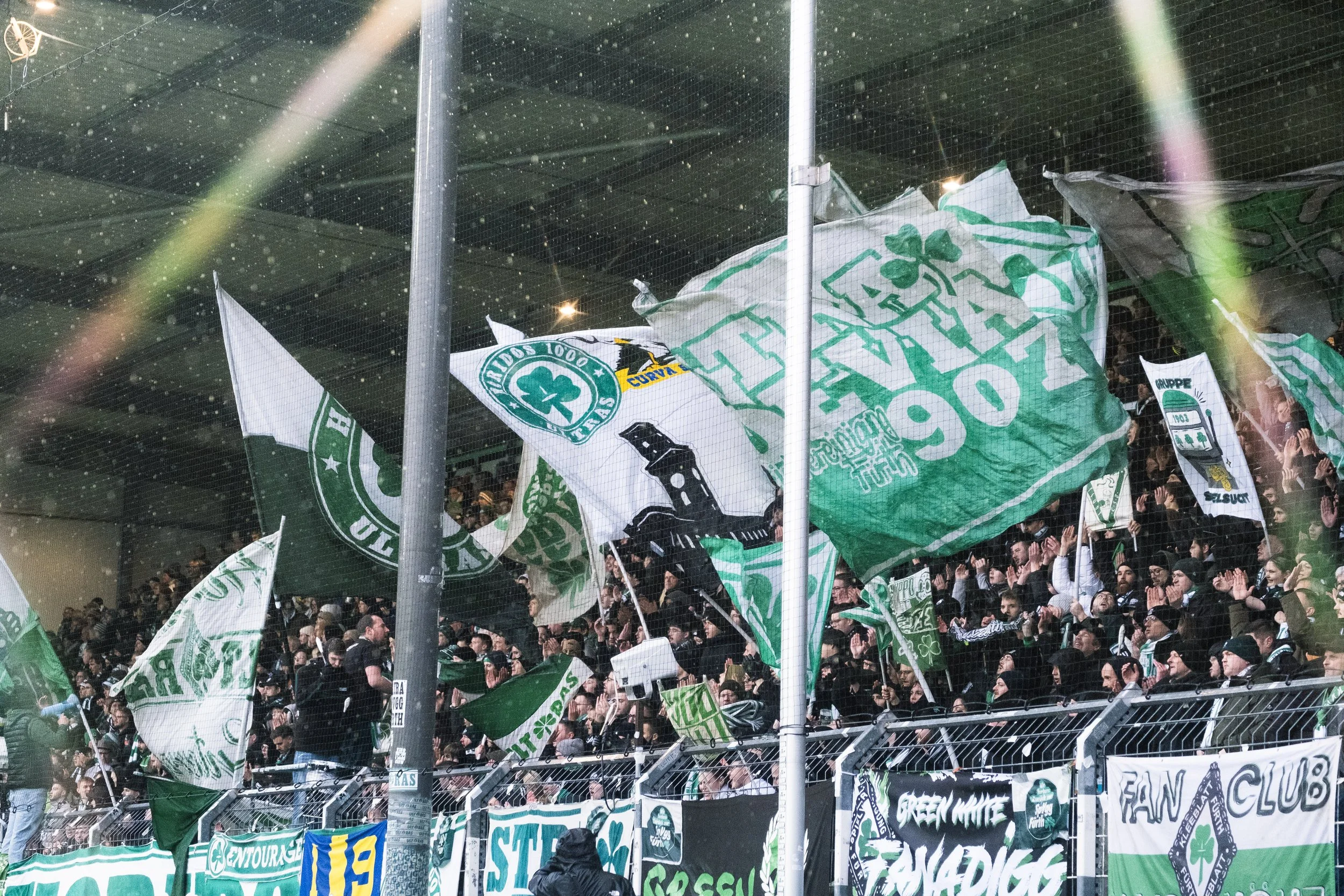 Menschenmenge im Stadion, Fans mit grün-weißen Fahnen, jubeln und klatschen, in der Kulisse eines Fußballspiels.