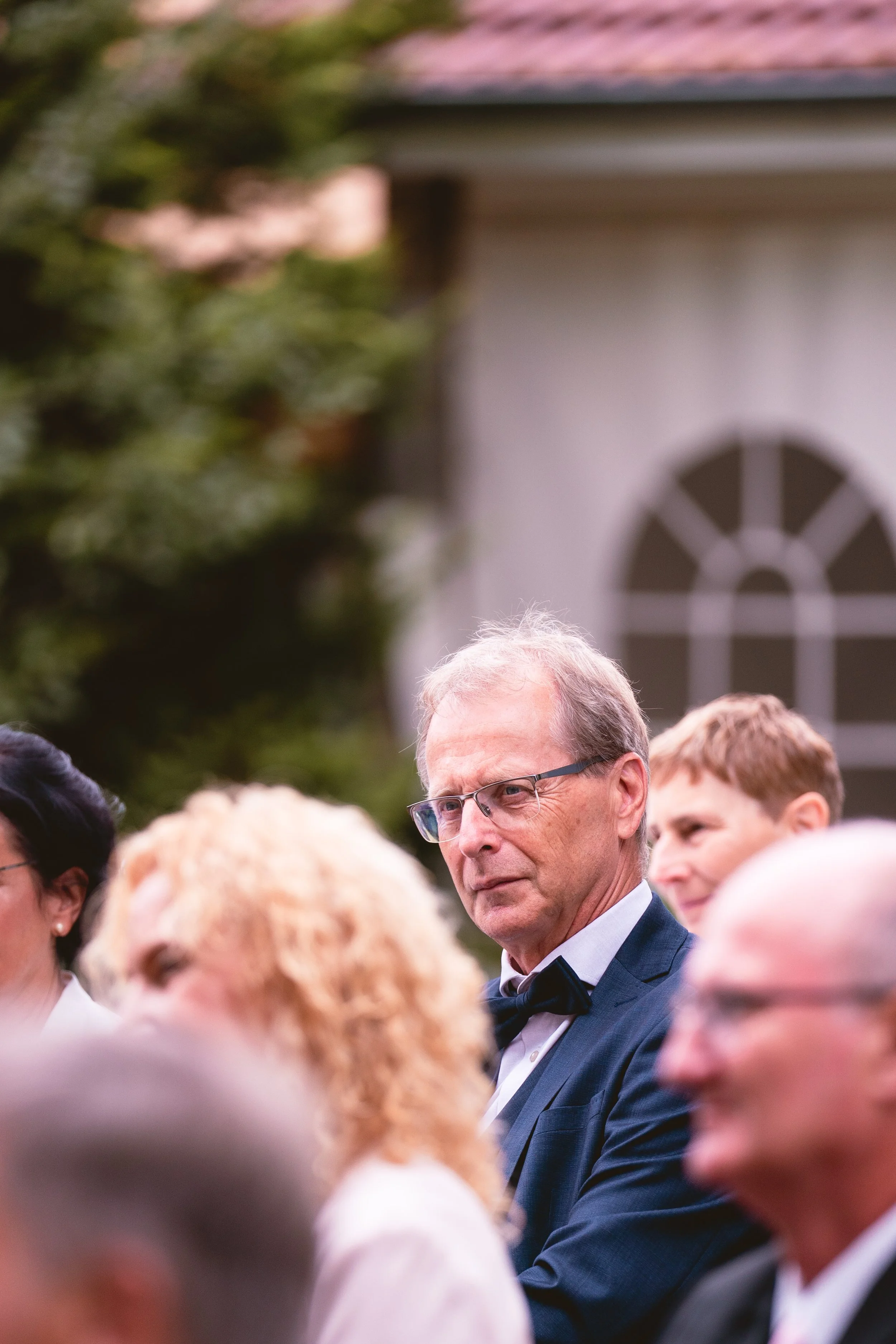 Ein Mann mit Brille in formeller Kleidung bei einer Veranstaltung, umgeben von mehreren Menschen im europäischen Stil, im Hintergrund ein Haus mit einer runden Fensteröffnung.