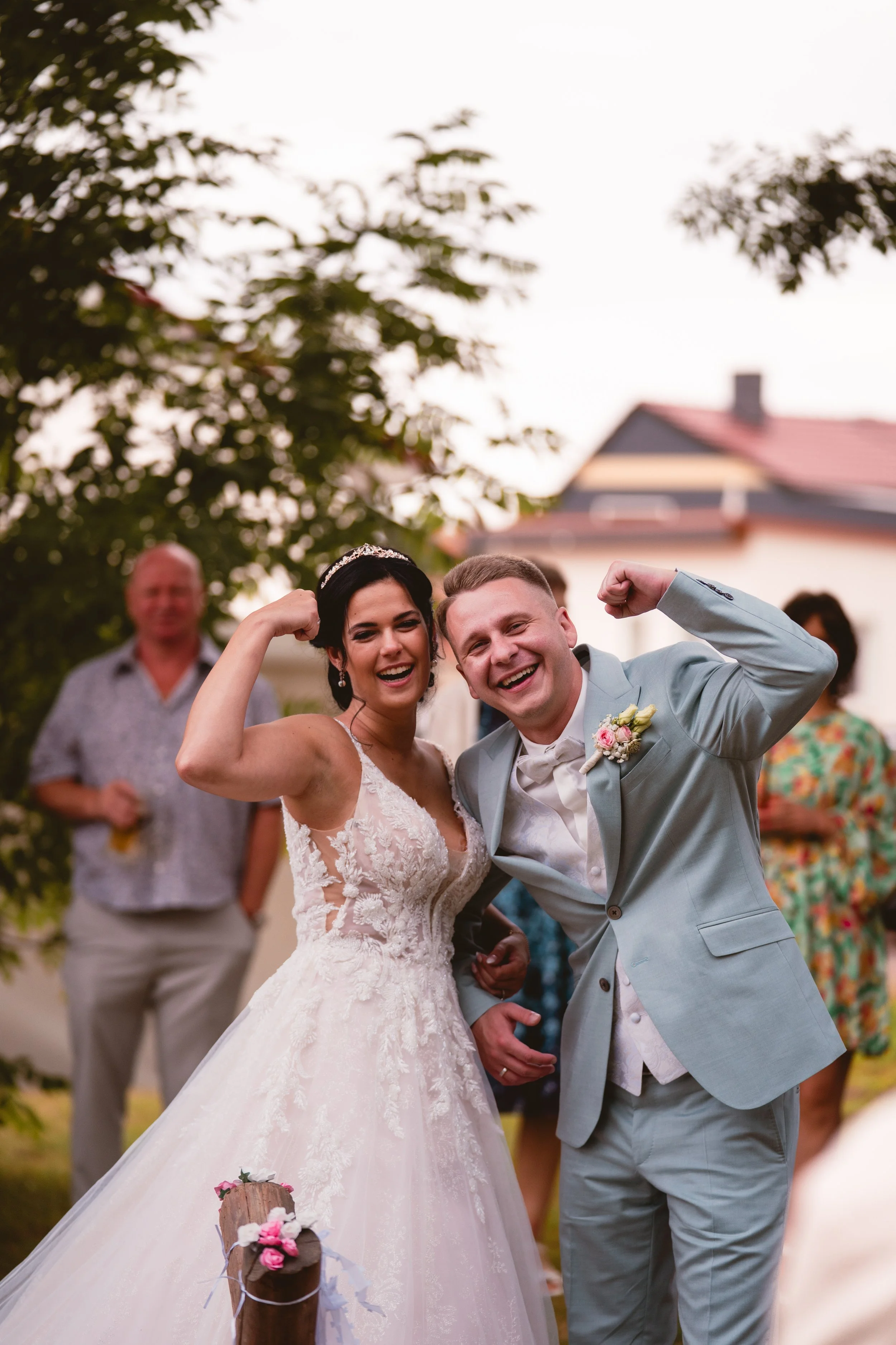 Ein glückliches Brautpaar, das bei ihrer Hochzeit in einem Garten feiert, beide tragen formelle Kleidung, sie ist in einem weißen Brautkleid und er in einem hellblauen Anzug, sie zeigen Muskeln und lachen, im Hintergrund sind Gäste und Bäume.