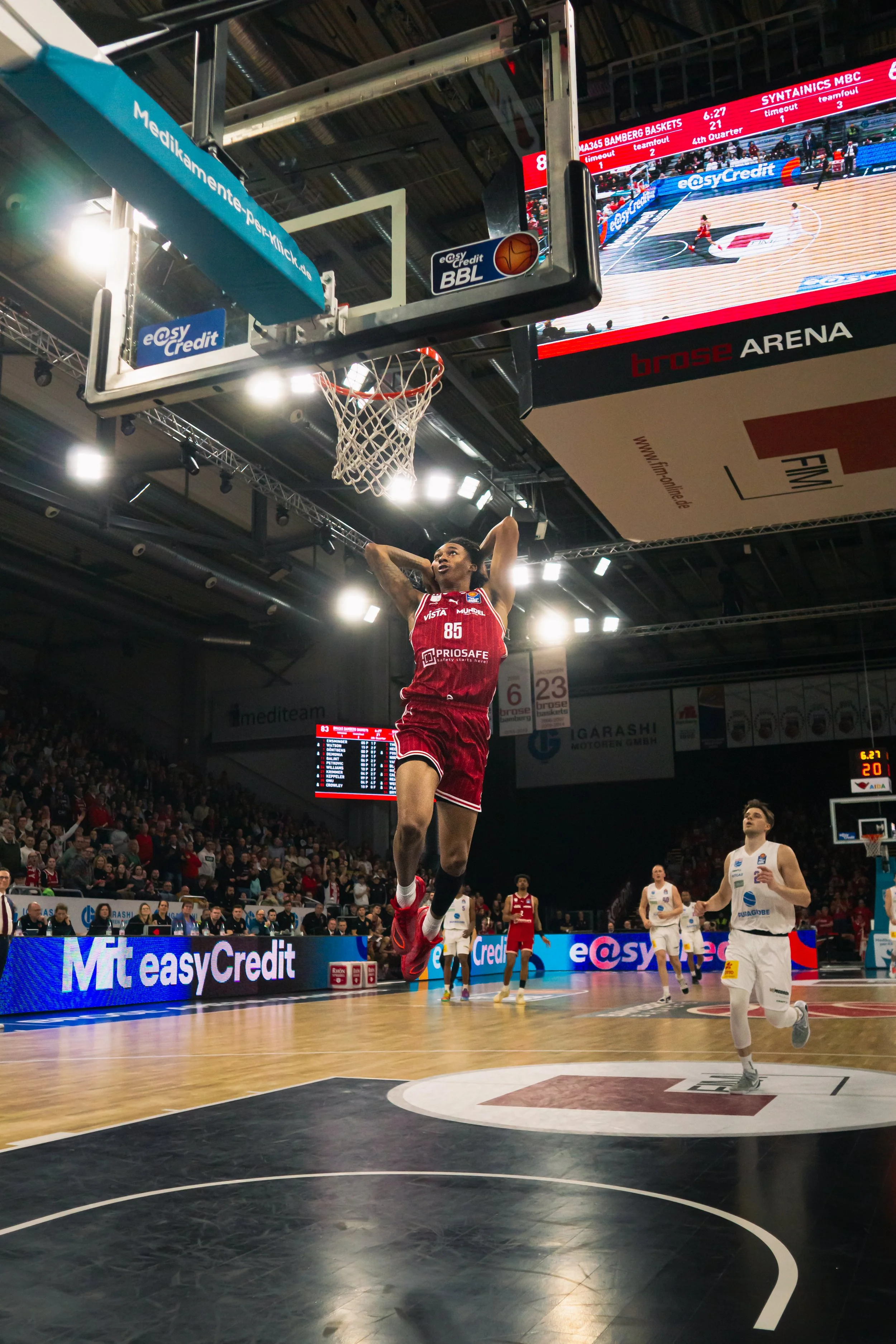 Ein Basketballspieler in rotem Trikot springt zum Korb, während andere Spieler im weißen Trikot auf dem Spielfeld stehen. Das Spiel findet in einer Halle mit Publikum statt, mit einem digitalen Anzeigetafel im Hintergrund.