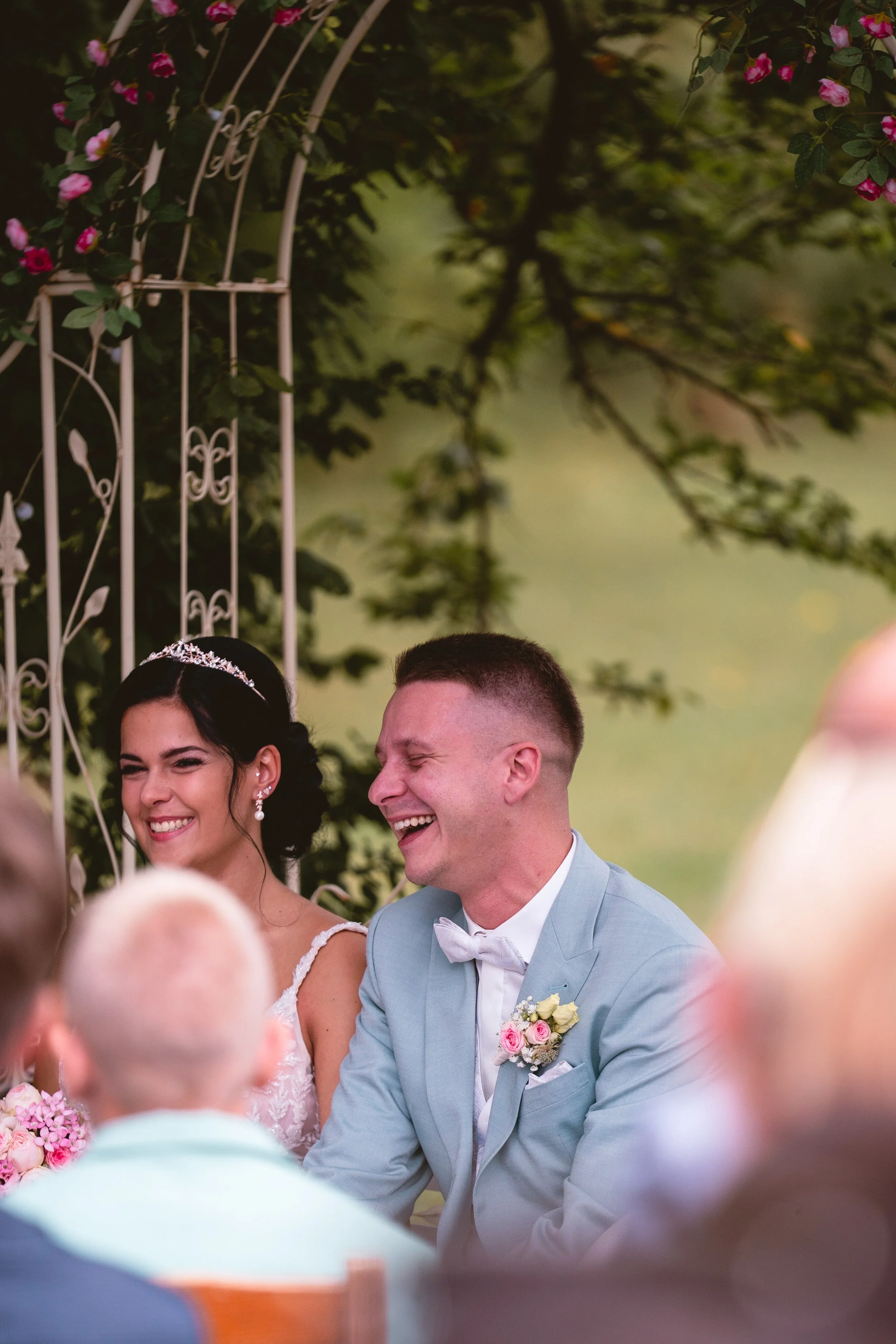 Ein lachendes Brautpaar bei einer Hochzeit, umgeben von Gästen, im Hintergrund grüne Bäume und ein weißer Gartenbogen mit rosa Blumen.