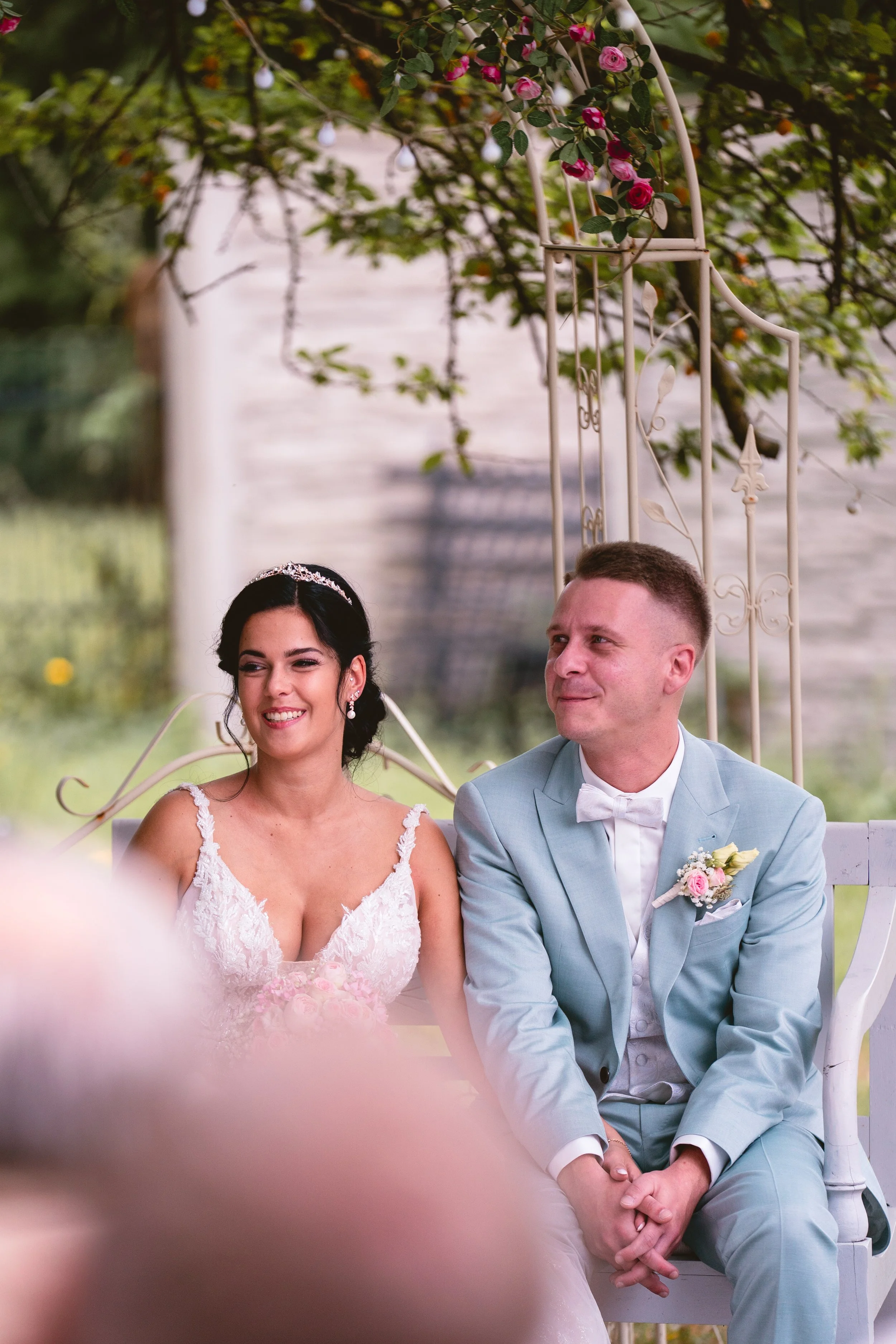 Ein Brautpaar sitzt im Garten bei einer Hochzeit. Die Braut trägt ein weißes Spitzenkleid mit Trägern und eine Tiara, der Bräutigam trägt einen hellblauen Anzug mit Fliege und eine Brosche. Über ihm hängt eine weiße Eisenbogen-Torso mit Rosen und grü